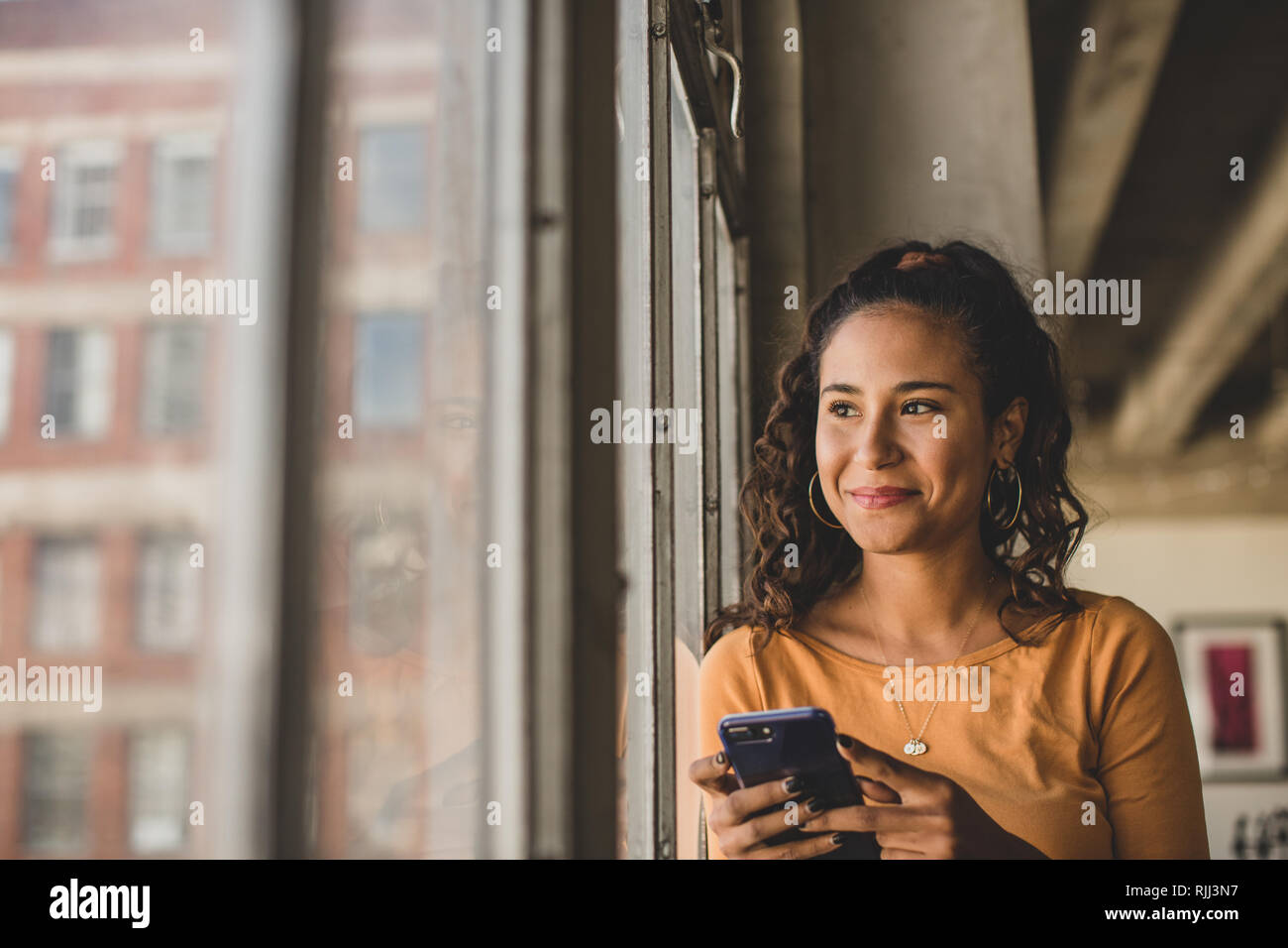 Junge Hispanic weiblichen Blick aus Fenster in loft apartment Holding smartphone Stockfoto