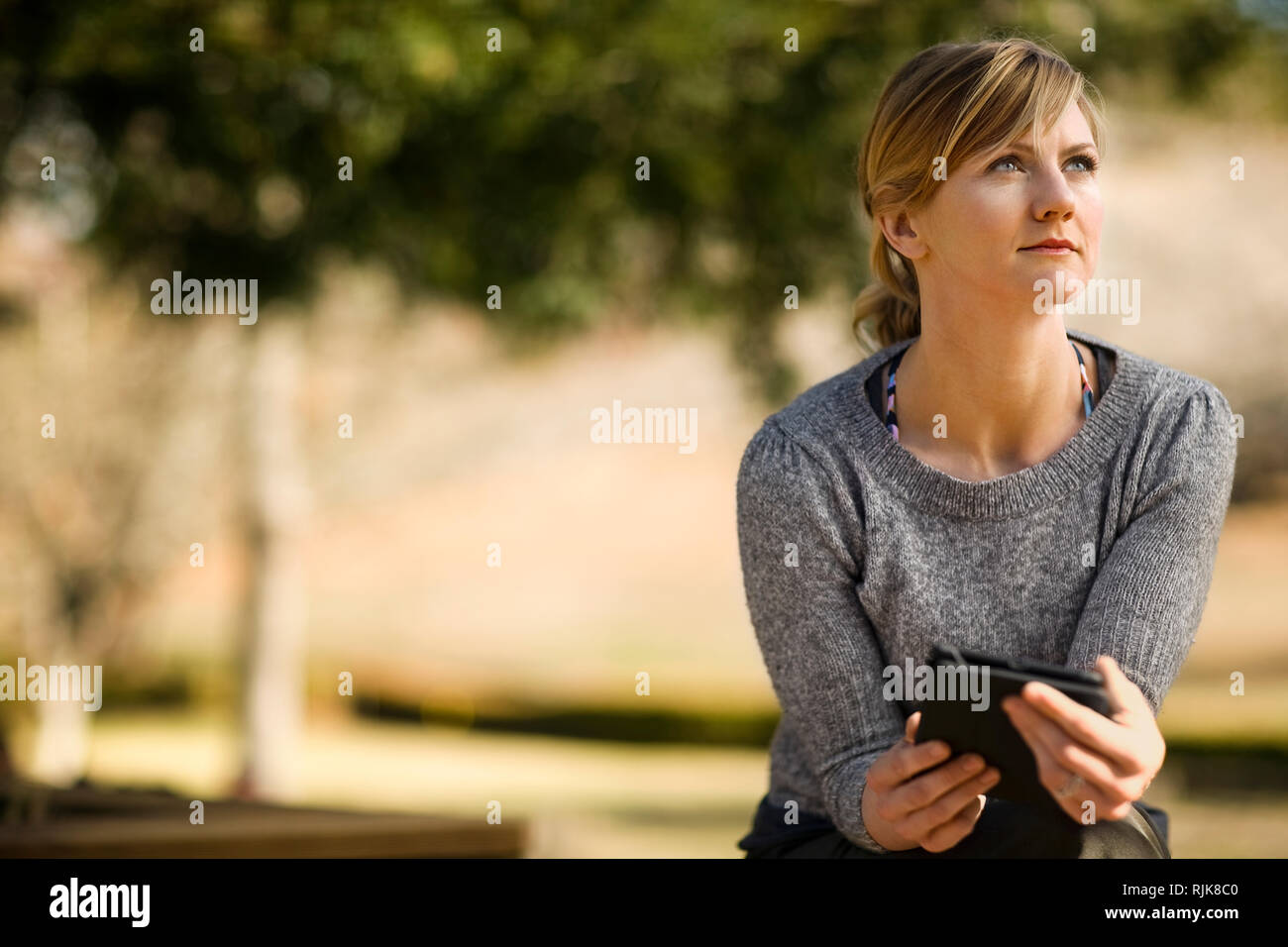 Junge Frau in einem sonnigen Garten sieht aus Ihrem digitalen Tablet und starrt nachdenklich in die Ferne. Stockfoto