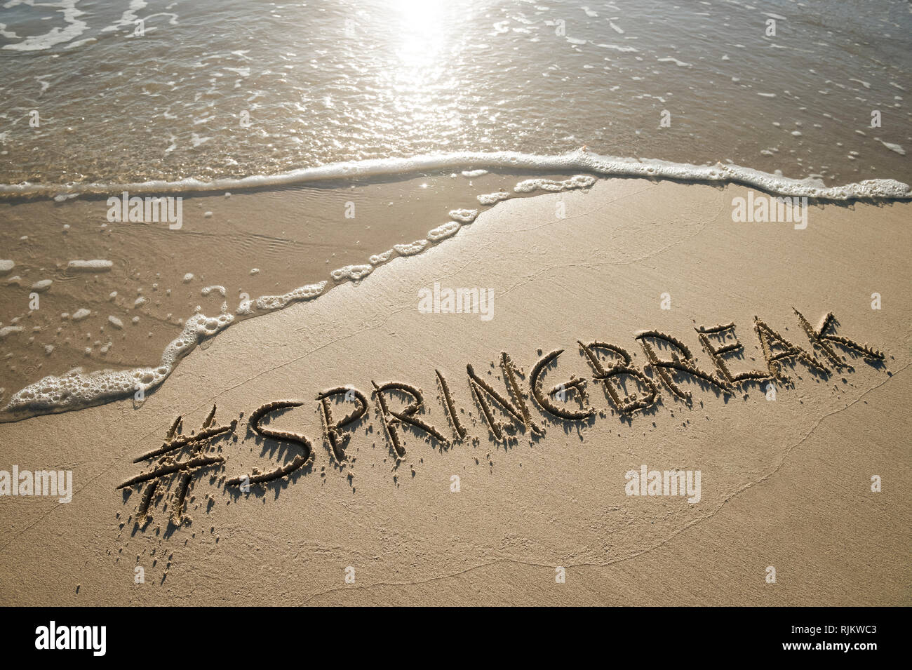 Moderne Spring Break Nachricht handschriftlich mit einem social media Hashtag auf der glatten Sand eines leeren Strand mit einem entgegenkommenden Wave Stockfoto