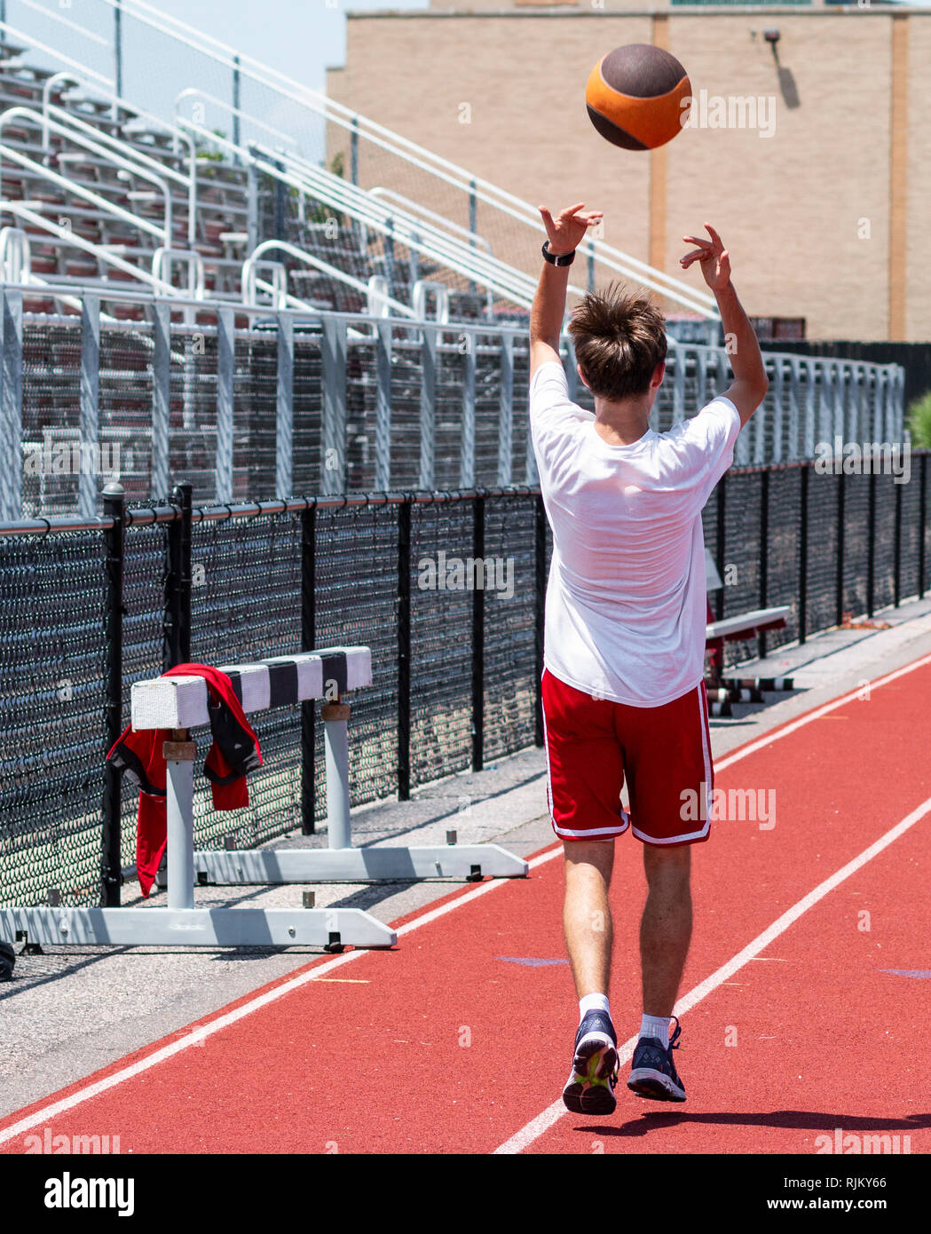 Ein High School Athlet ist auf einer roten Piste vor der Bleiche werfen einen Medizinball overhead rückwärts für Kraft, Schnelligkeit und Beweglichkeit zu üben. Stockfoto