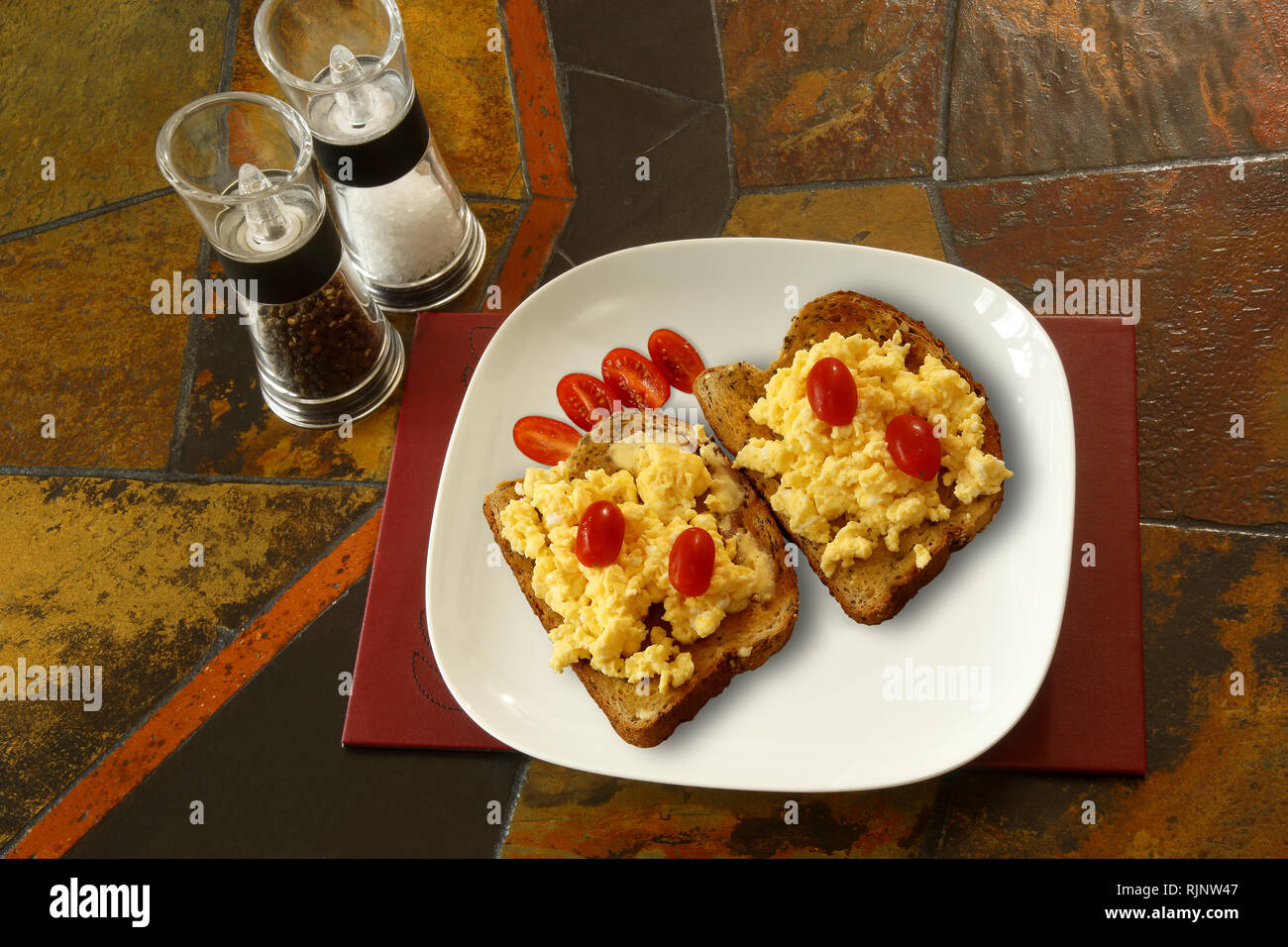 Rühreier auf Toast mit Pflaumentomaten. Hintergrund ist eine geflieste Tischplatte und zeigt Salz- und Pfeffermühlen Stockfoto