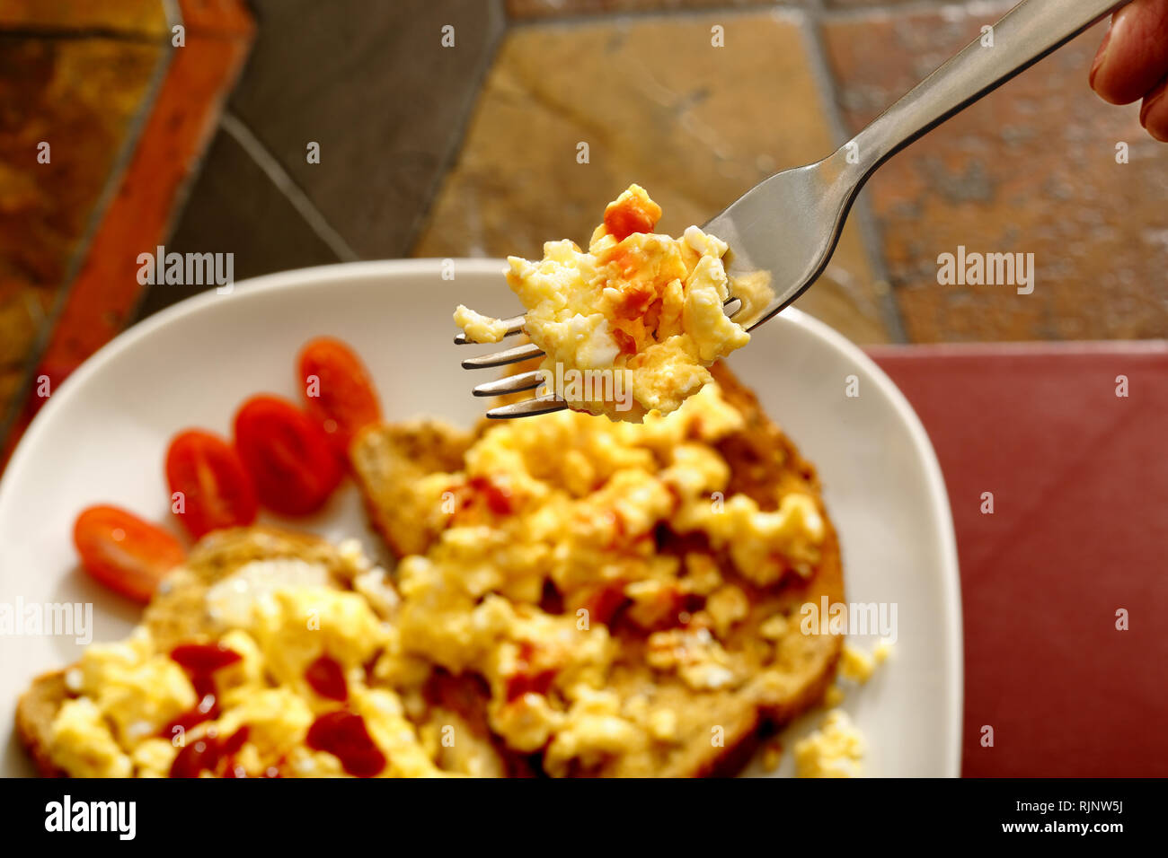 Rührei auf Toast mit Pflaumentomaten und Tomatensauce. Hintergrund ist eine Kachel-Tischplatte. Stockfoto