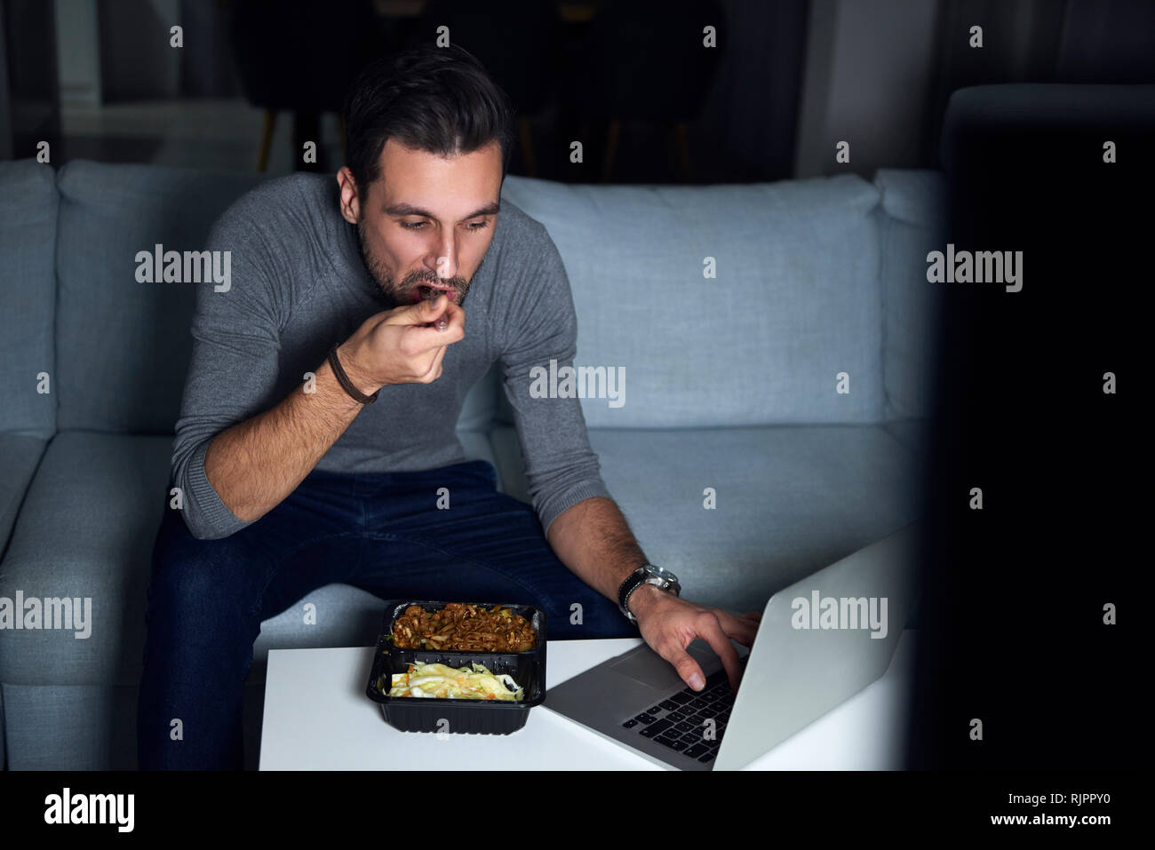 Junge Mann sitzt auf einem Sofa in Abend Essen zum Mitnehmen und mit Laptop Stockfoto
