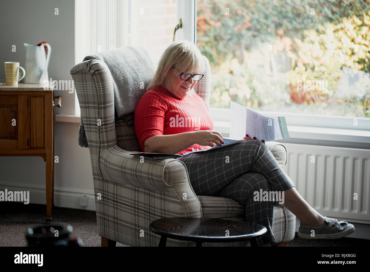 Eine ältere Frau sitzt in einem Sessel im Wohnzimmer ihres Hauses, Lesen durch Schreiben. Stockfoto