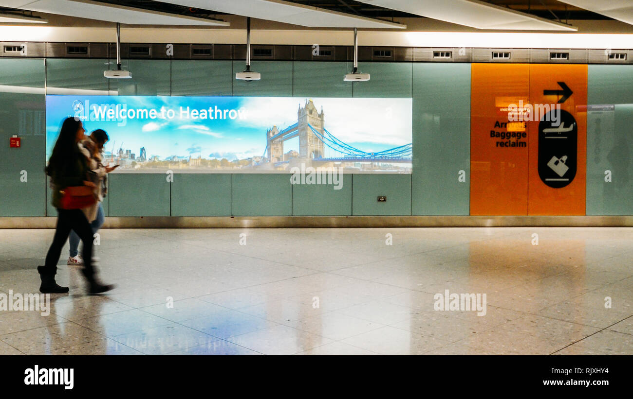 London Heathrow, UK, Feb 5, 2019: Zwei Passagiere vorbei gehen. Ein Zeichen und Bild der Tower Bridge geschrieben Willkommen zum Flughafen Heathrow Stockfoto
