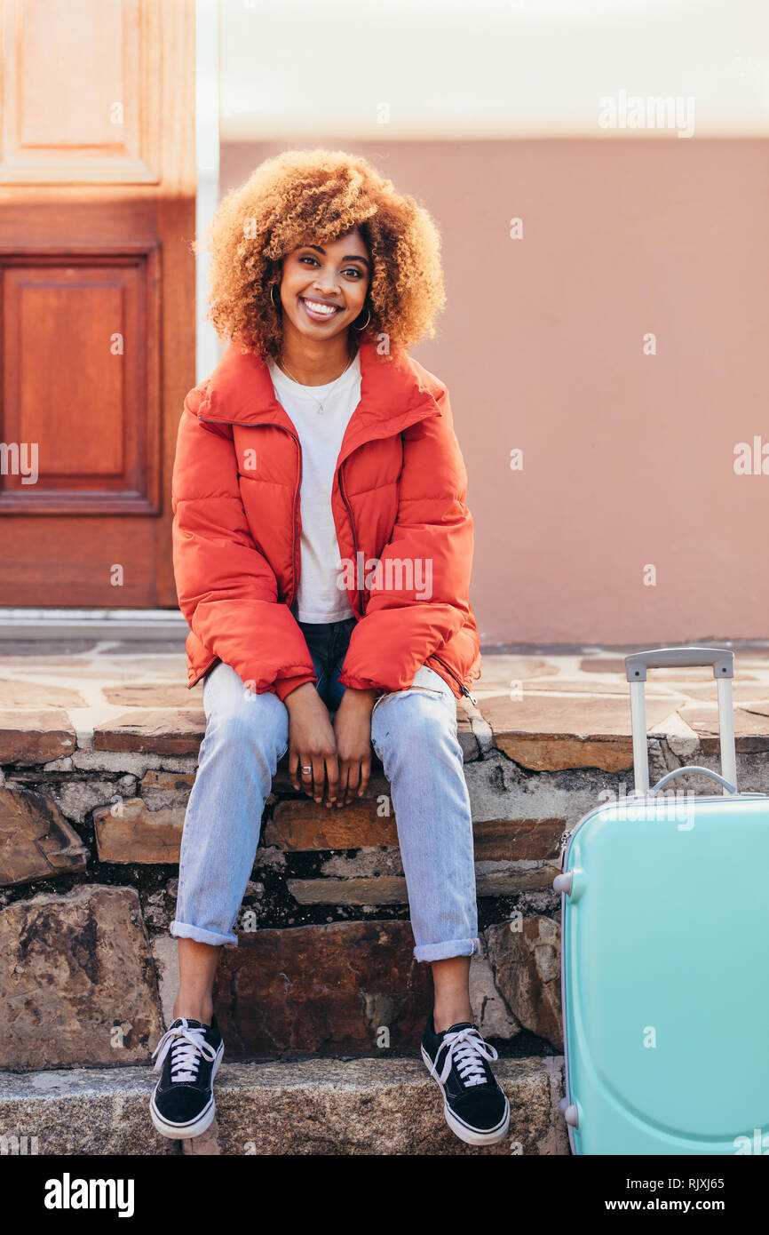 Lächelnd afro-amerikanische weibliche Reisende im Urlaub außerhalb eines Hauses mit ihrem Gepäck Tasche sitzen. Lächelnde Frau auf einen Urlaub im Freien sitzen, mit einem tr Stockfoto
