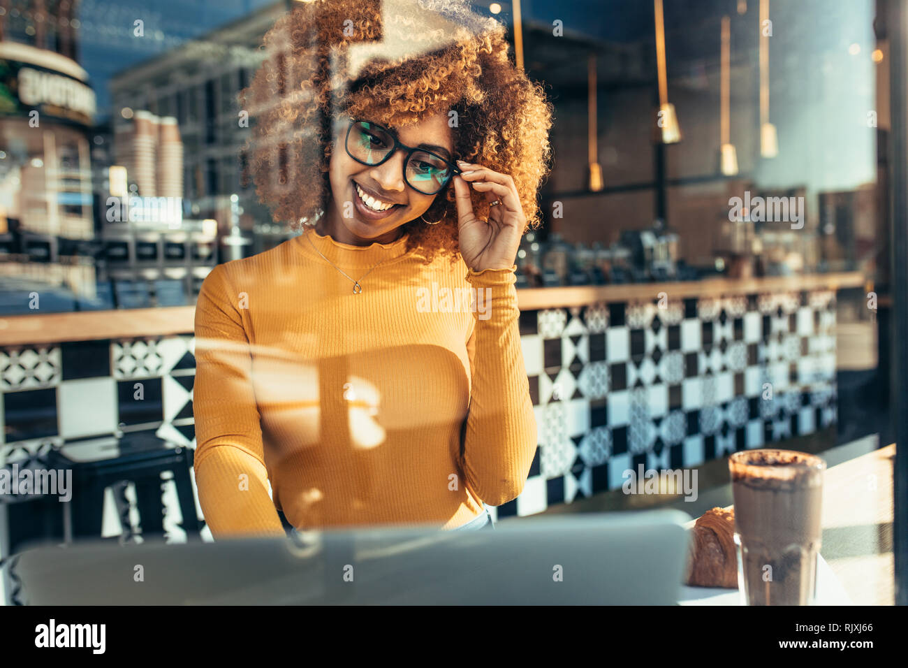 Lächelnd afro-amerikanische Frau, die Arbeiten am Laptop in einem Café sitzen. Fröhliche Frau in Brillen sitzen in einem Restaurant an einem Laptop gesehen Stockfoto