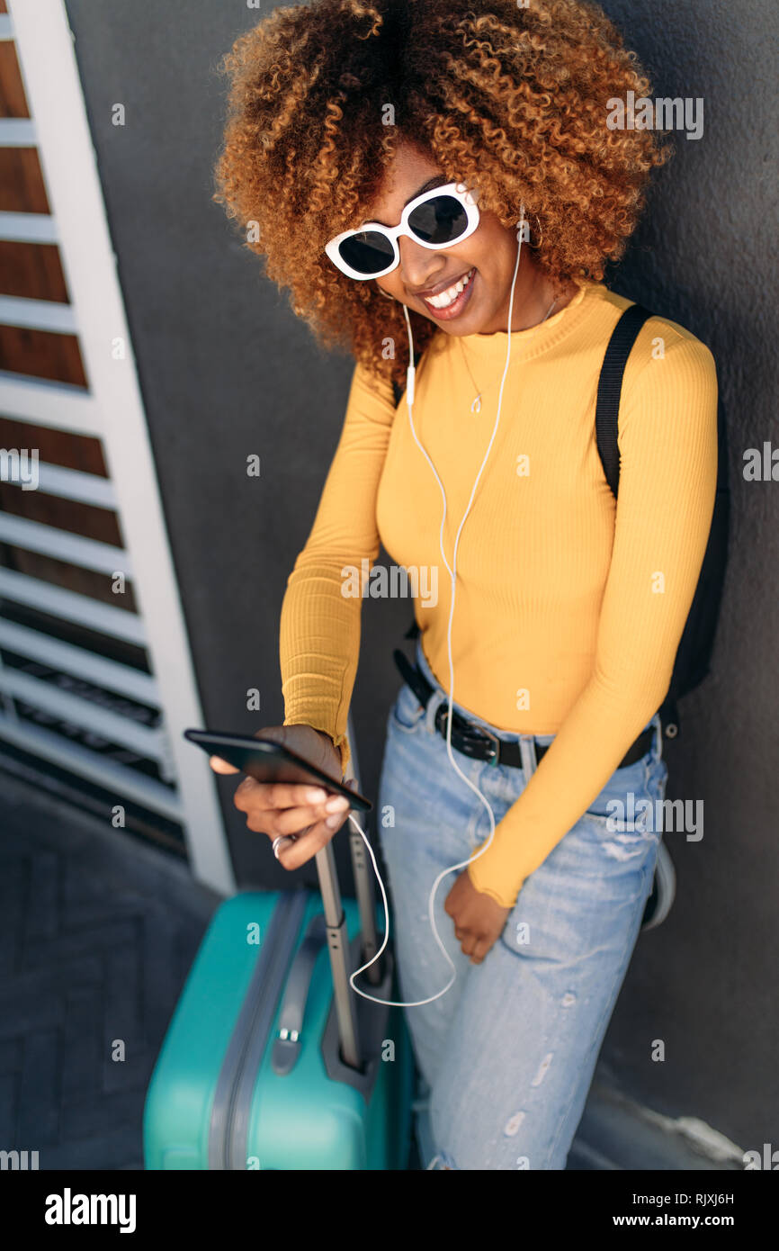Lächelnd afro-amerikanischen Touristen stehende Frau mit ihrem Gepäck mit Handy in der Hand. Fröhliche Frau Reisenden in Sonnenbrille hören musi Stockfoto