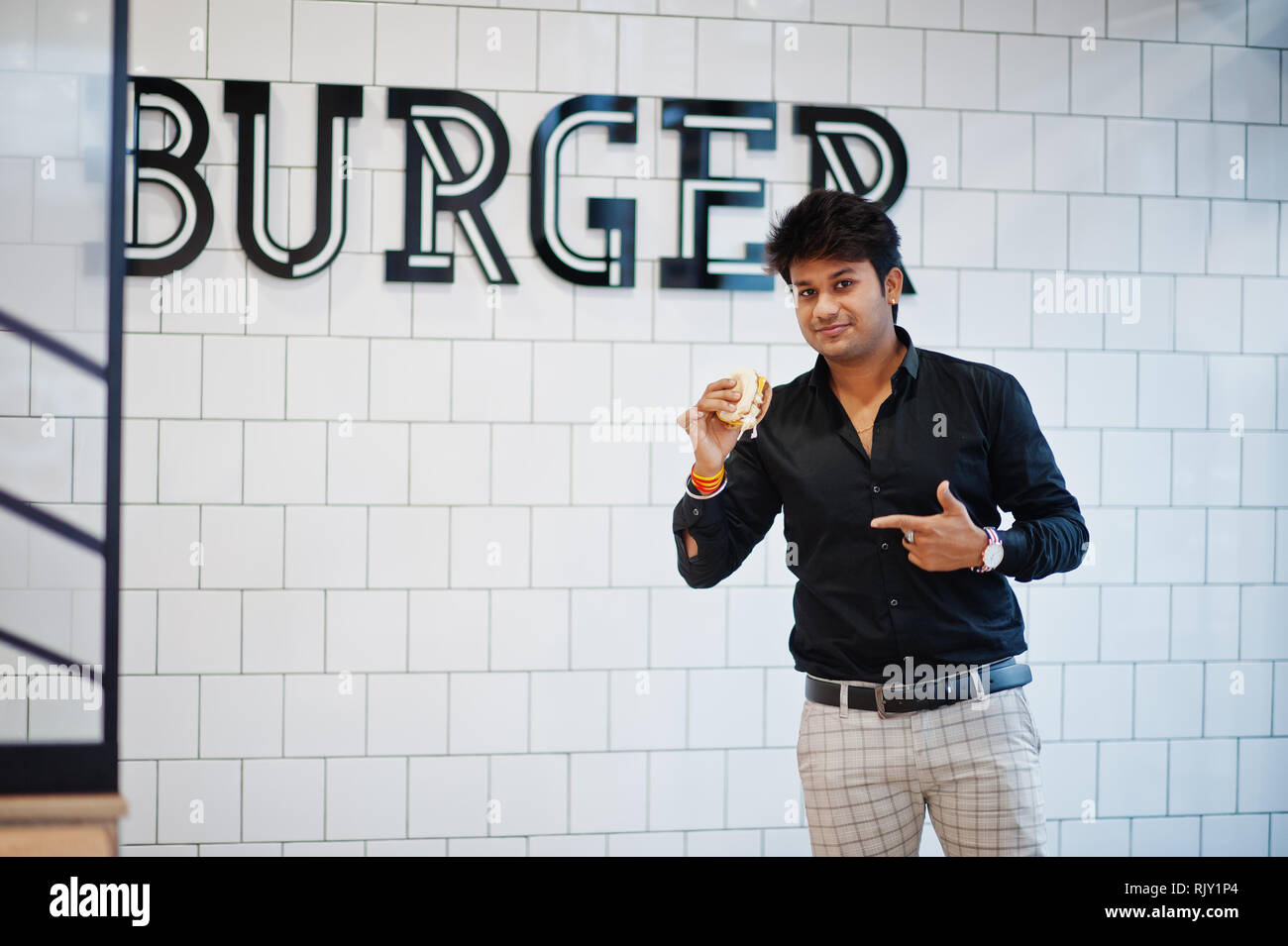 Stilvolle Inder in Fast-food Cafe mit Hamburger in der Hand gegen Burger Zeichen an der Wand. Stockfoto