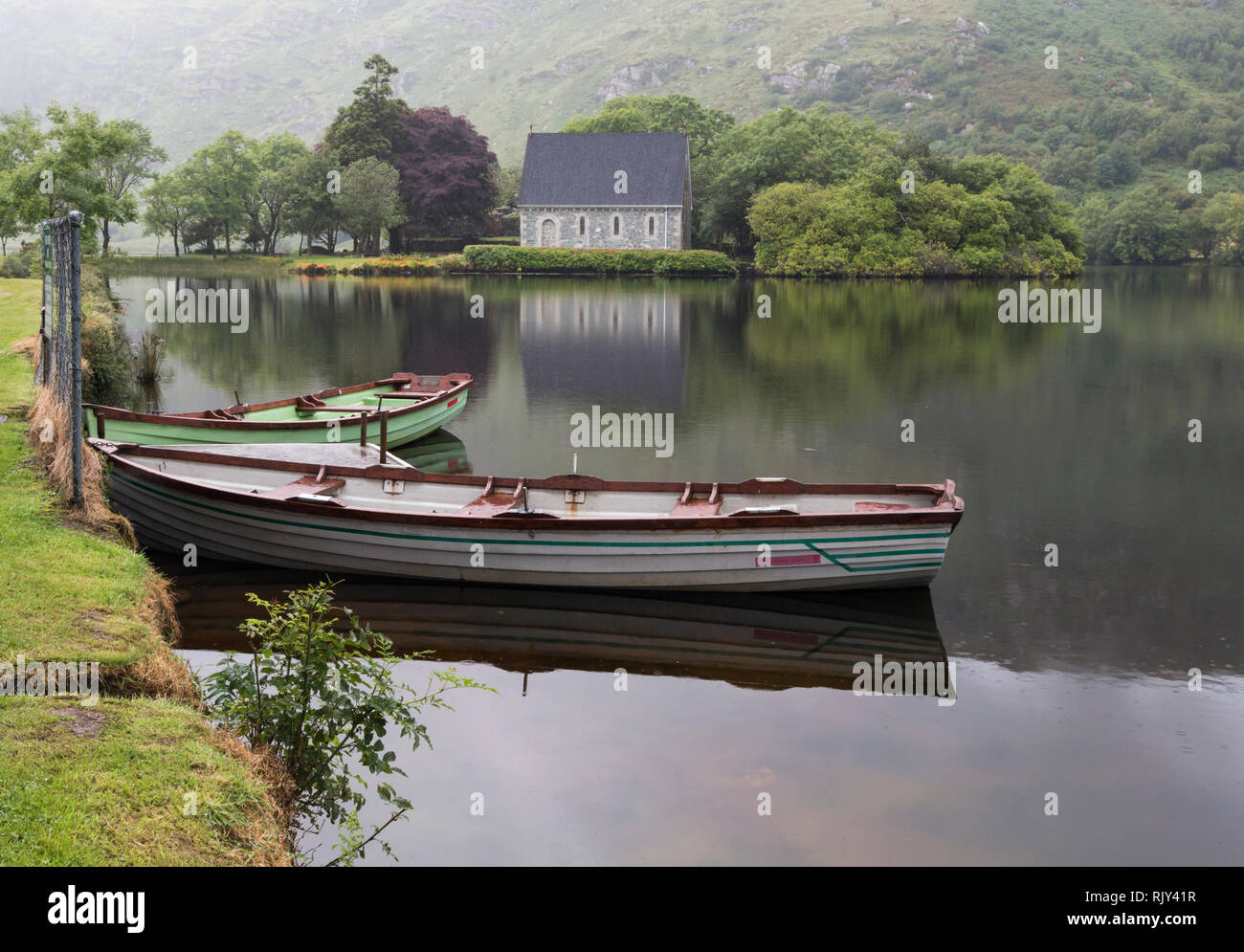 Die St. Finbarr Oratorium über den See bei Gougane Barra, County Cork, Republik Irland gesehen. Eire. Ruderboote im Vordergrund. Stockfoto