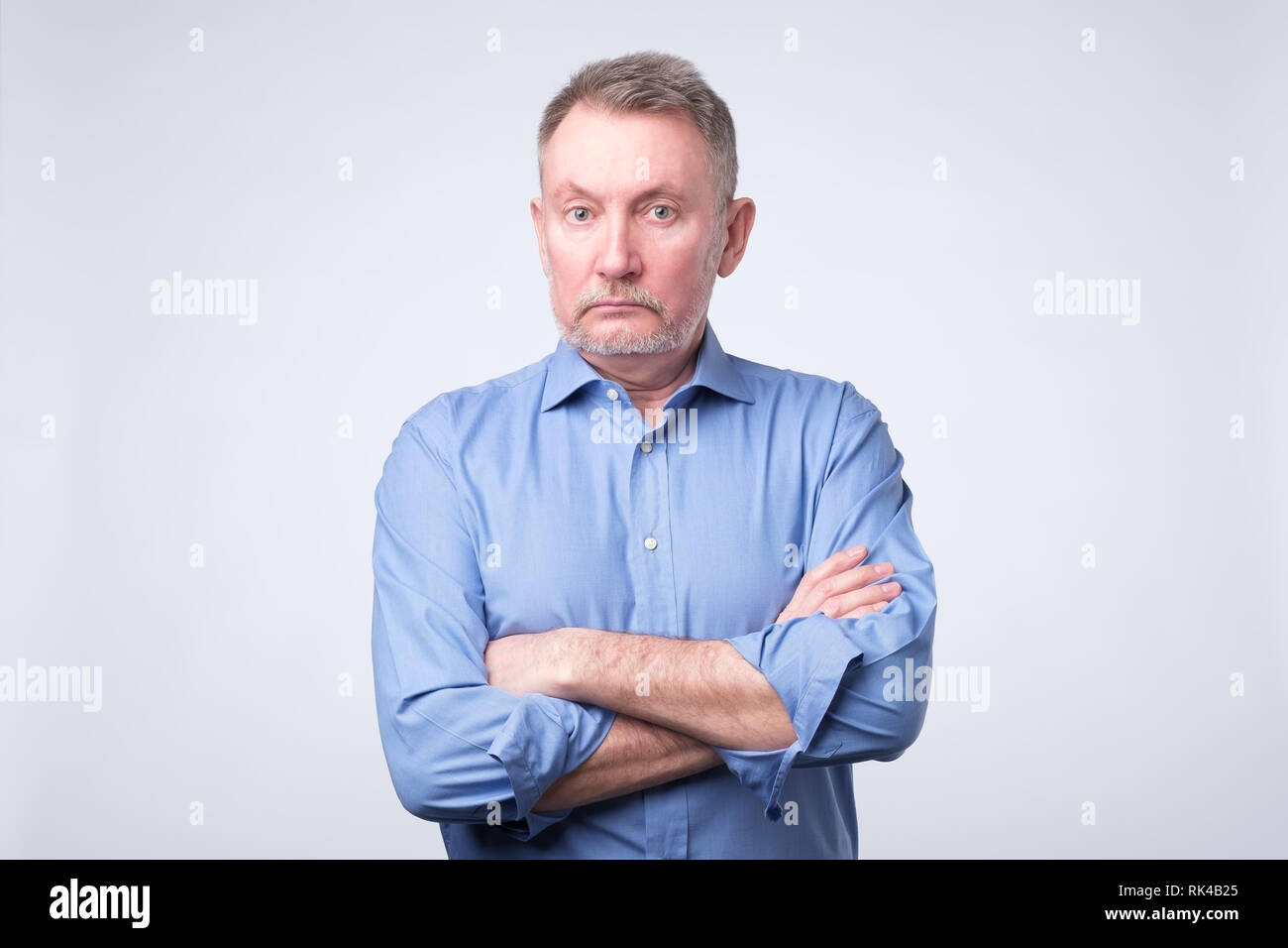 Ernsthafte älterer Mann mit verschränkten Armen und einem ausdruckslosen Ausdrucks auf graue Wand posieren Stockfoto