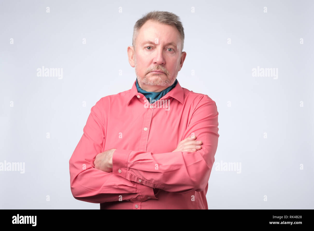 Ernsthafte älterer Mann mit verschränkten Armen und einem ausdruckslosen Ausdrucks auf graue Wand posieren Stockfoto