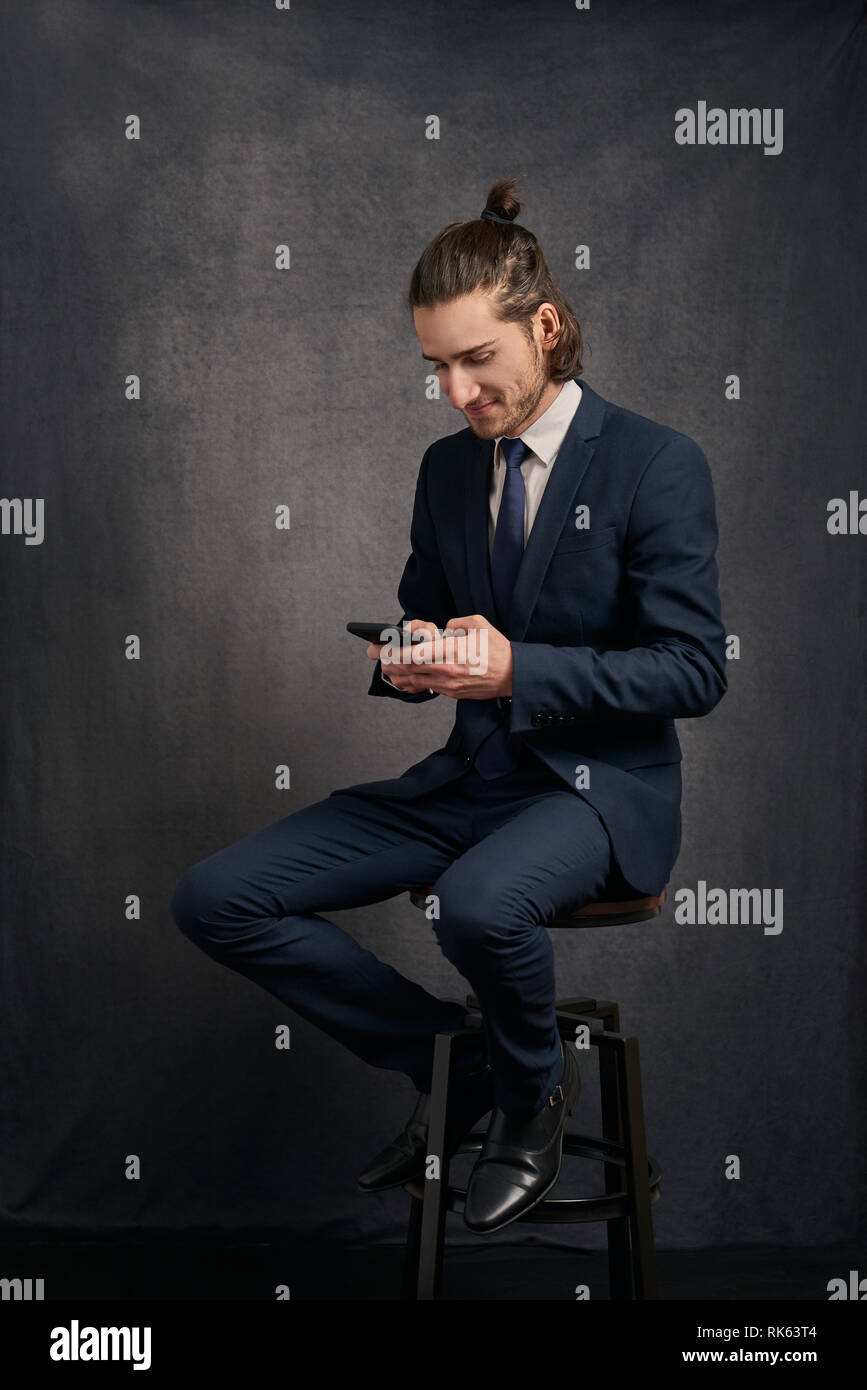 Stilvolle hübscher junger Mann mit langen Haaren in einem Brötchen, trägt Anzug, sitzen auf den Barhocker und Smartphone in seinen Händen, Simsen. Stockfoto