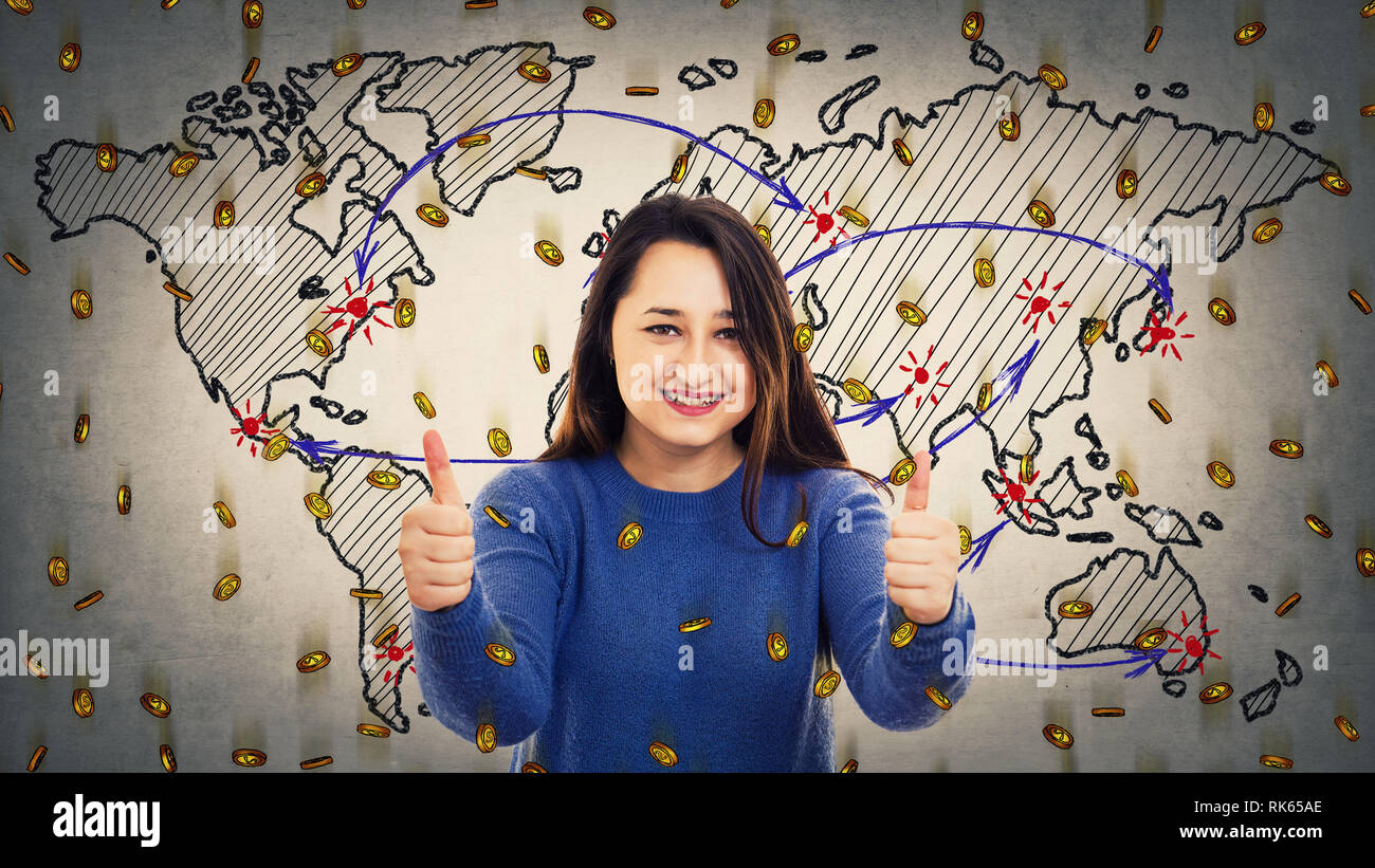 Fröhliche junge Frau, Daumen hoch positive Geste, wie Zeichen über Weltkarte Hintergrund und viele goldene Münzen nach unten fallen. Global cu Stockfoto