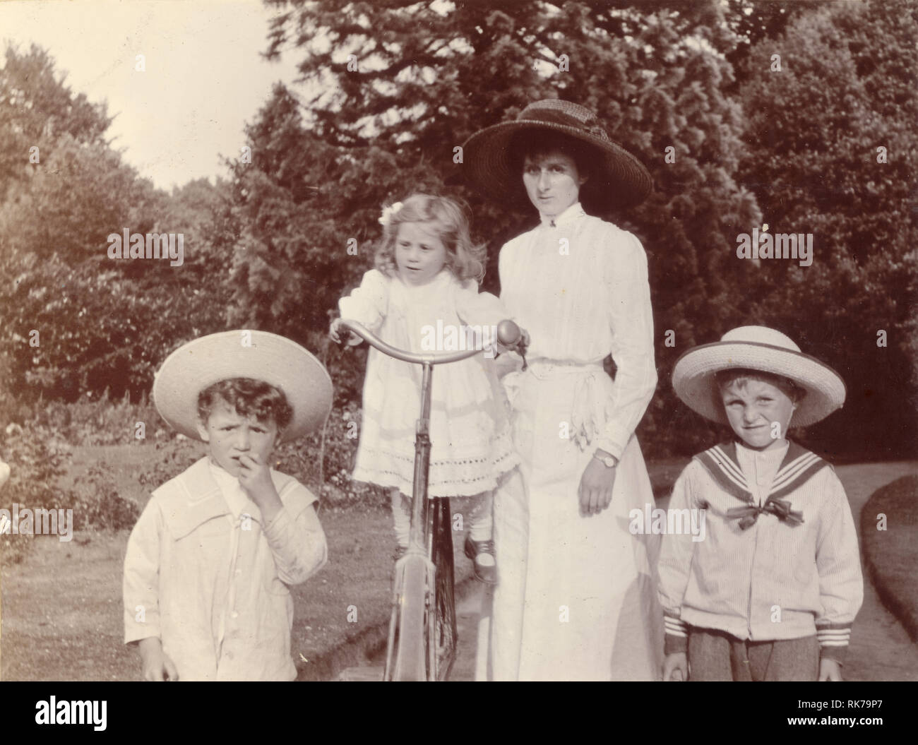 Original Vintage Cycling Edwardian Gruppenfoto einer attraktiven Mutter in einer weißen Sommerbluse und einem Rock, die mit ihren Kindern im Park oder Garten spazieren geht. Das jüngste Kind ist auf dem Fahrrad der Mutter ausgeglichen, die Jungs tragen modische Matrosenanzüge und Strohhüte, ca. 1910, Großbritannien Stockfoto