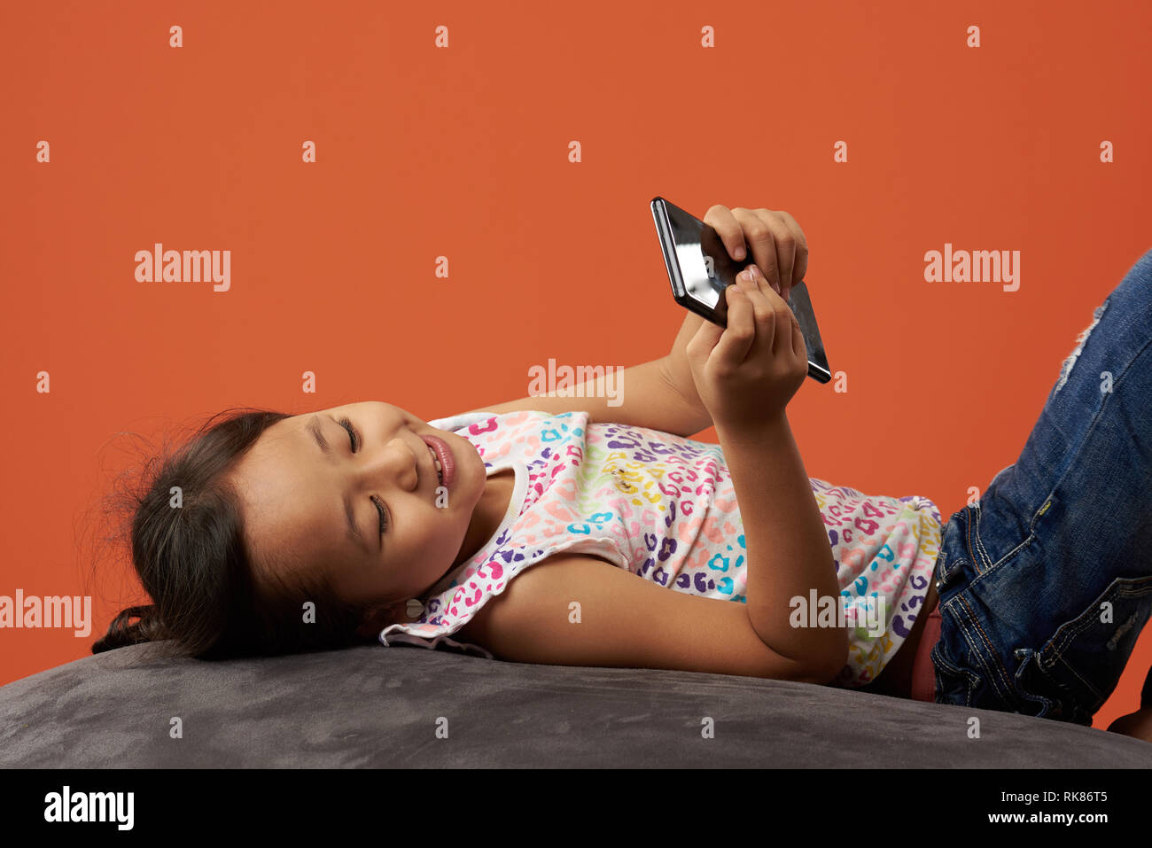 Kind lächelnd auf einem Sitzsack holding Telefon. Asiatische Mädchen, dass Handy und glücklich zu sein. Stockfoto