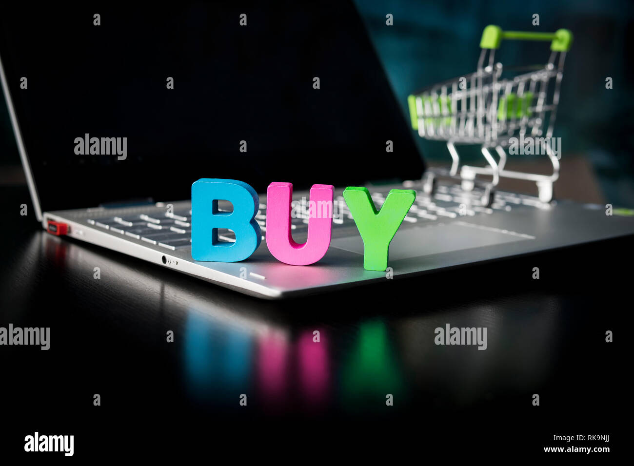 Kreative Schuß eines Holz- viele - farbige Wort kaufen auf Laptop unten an der schwarzen Bildschirm und Spielzeug Einkaufswagen Hintergrund. Konzept der kaufen Stockfoto