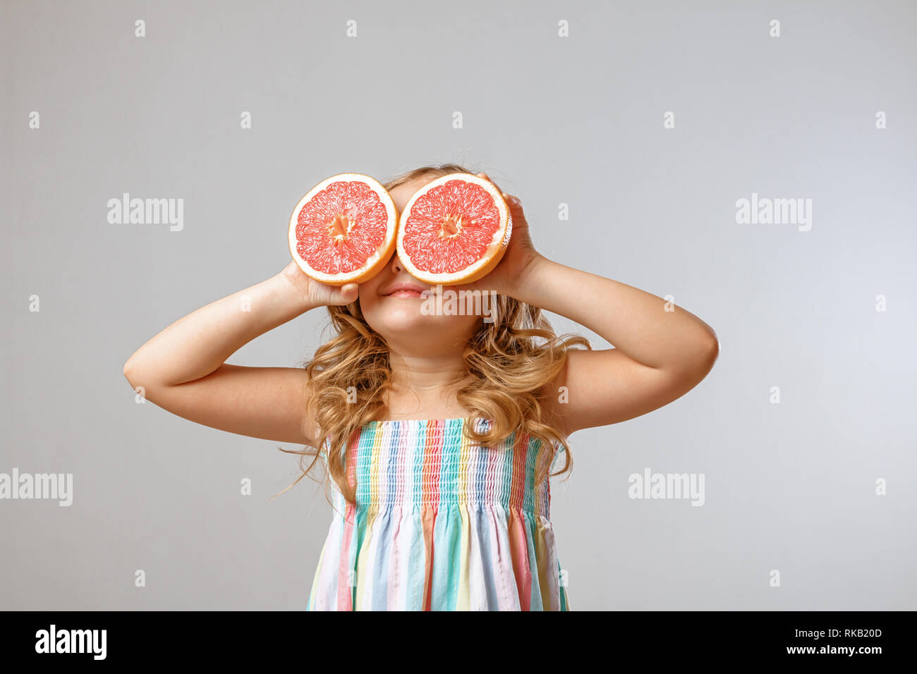 Fröhliche kleine Mädchen ist, das Spaß und Substitute Hälften Grapefruit anstelle der Augen. Grauer Hintergrund, Studio Stockfoto
