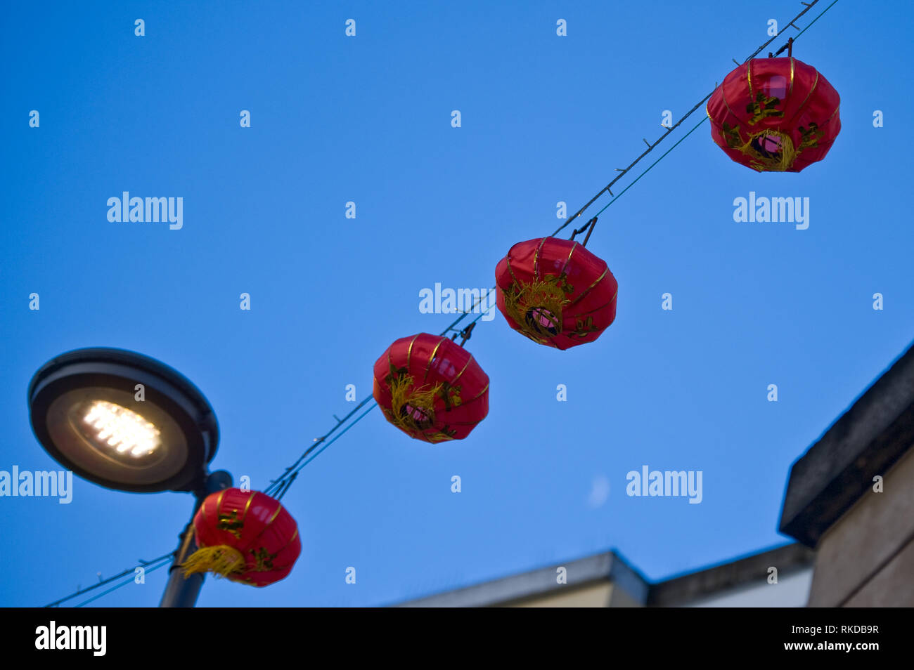 Stock Bild von vier roten Lampions hängen Kabel über einen Laternenpfahl vor blauem Himmel mit Mond leicht sichtbar unten rechts. Stockfoto
