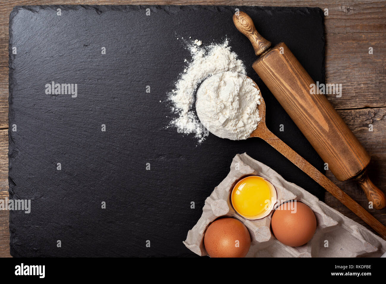 Zutaten und Utensilien zum Backen. Löffel mit Mehl, Eiern, Nudelholz auf schwarzem Schiefer Hintergrund. Draufsicht, Tiefenschärfe, Textfreiraum Stockfoto