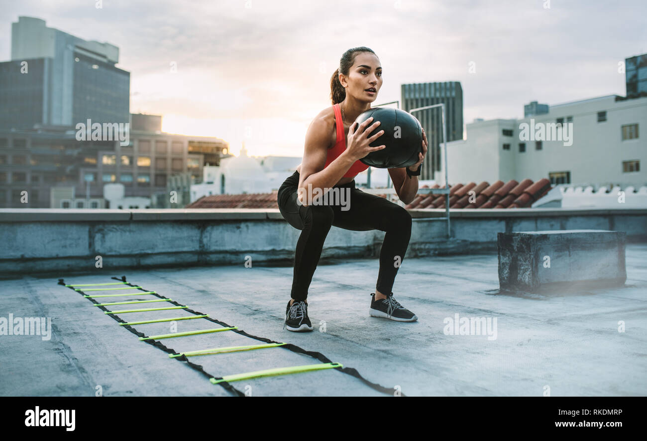 Weibliche Athleten dabei hockt Halte einen Medizinball auf einem Dach. Frau tun Trainings mit Medizinball mit einer Agilität Leiter durch ihre Seite Stockfoto