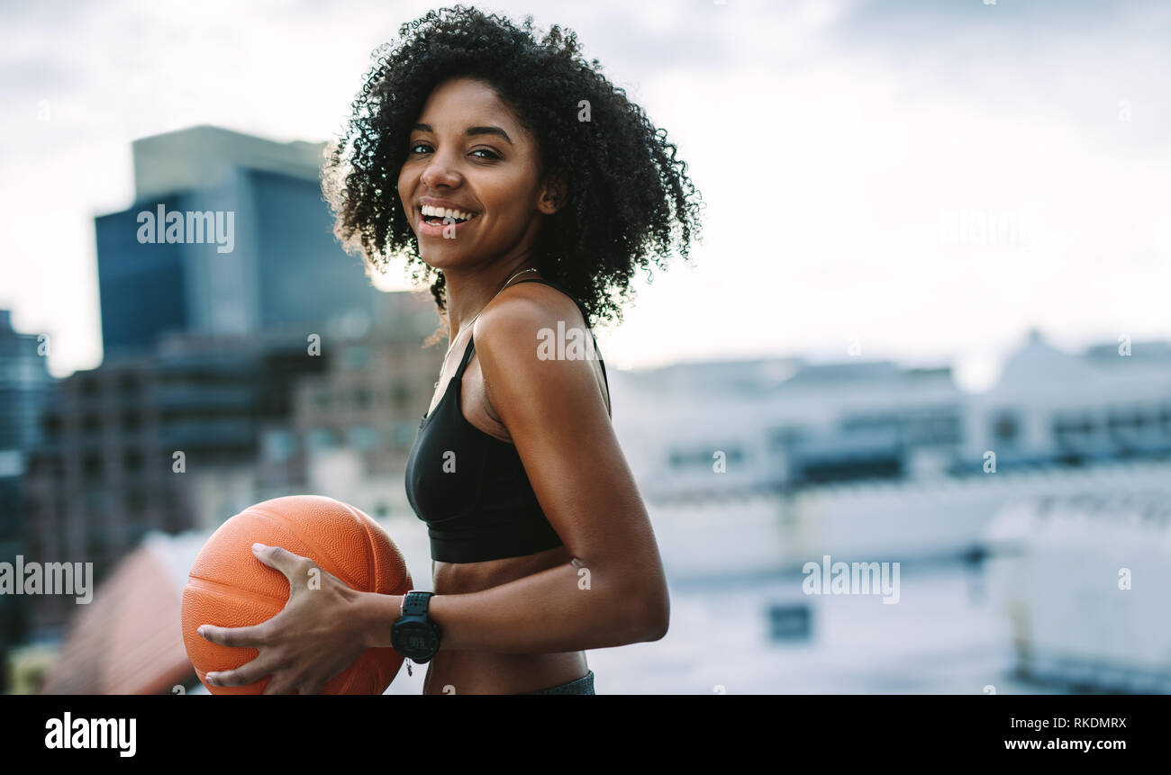 Seitenansicht Porträt eines lächelnden Frau Athlet mit einem Basketball stehen auf dem Dach. Frau in Fitness verschleiß Training mit einer Kugel auf dem Dach. Stockfoto