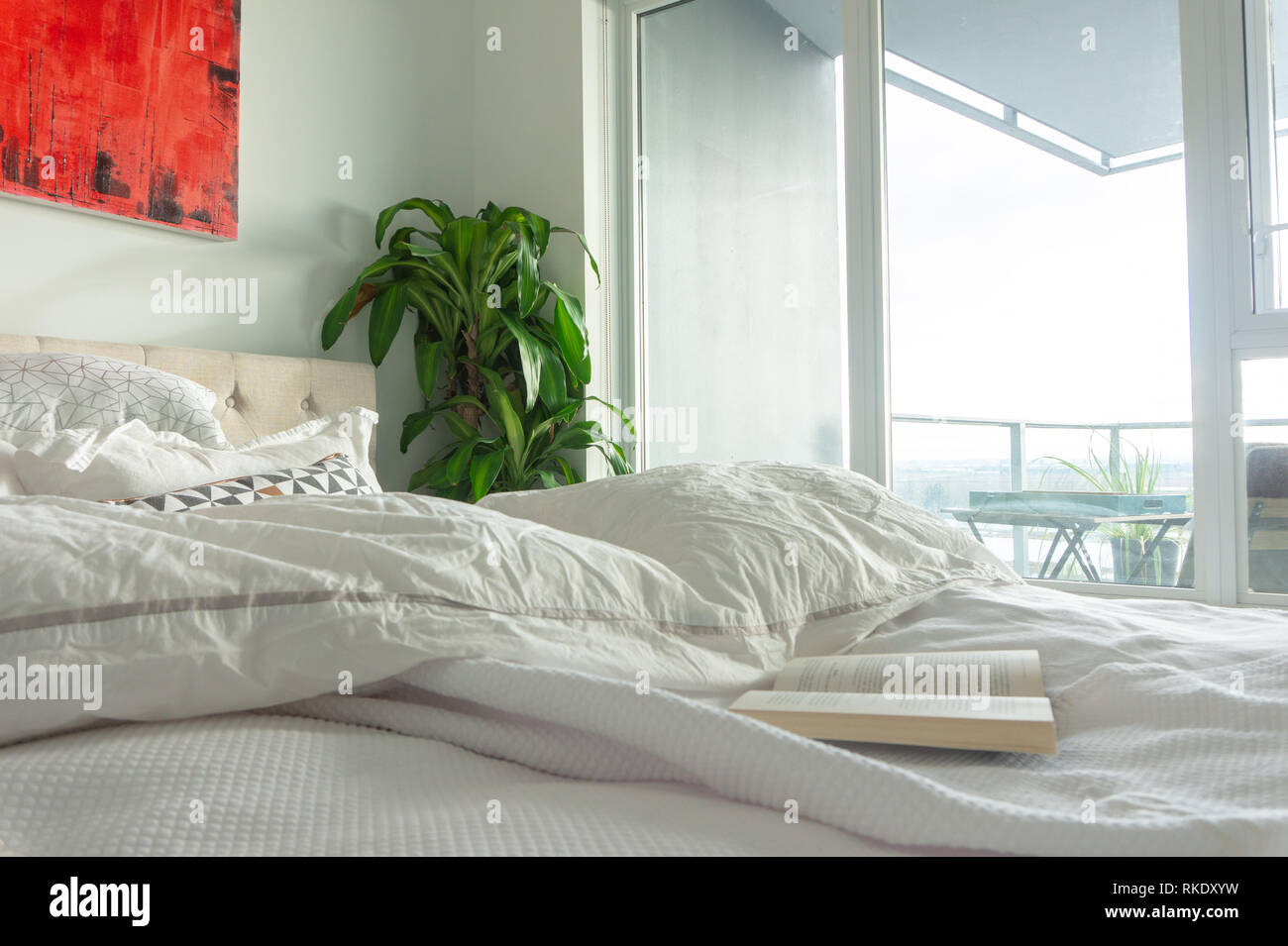 Buch über zerknittert, gebraucht Bett mit Wand Kunst in ein helles Apartment oder vrbo, Schlafzimmer mit großen Fenstern und Fenster Licht. Darstellung einer staycation, entspannend. Stockfoto