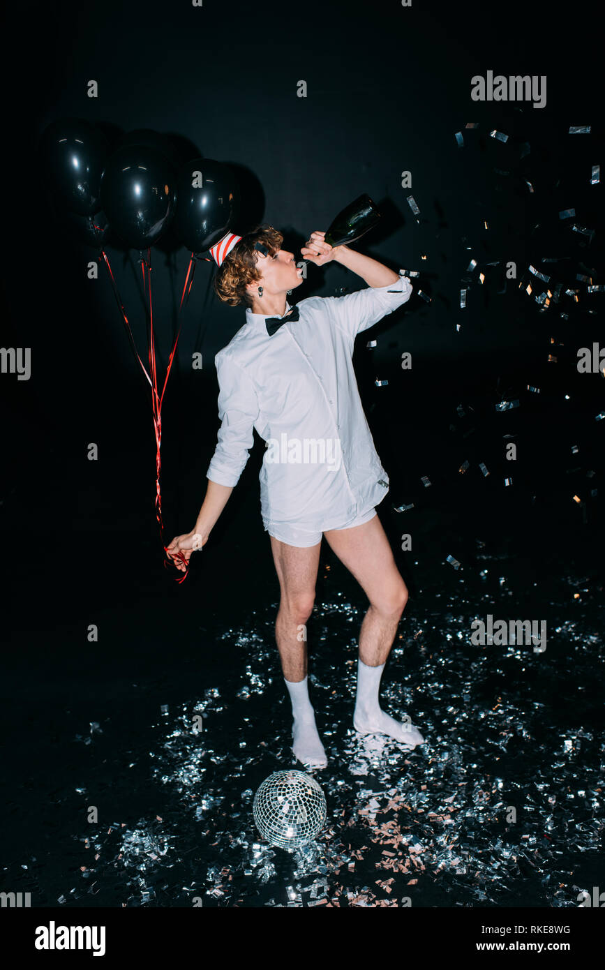 Mann mit lockigem Haar trinken Sekt aus der Flasche und Holding Ballons isoliert auf Schwarz Stockfoto