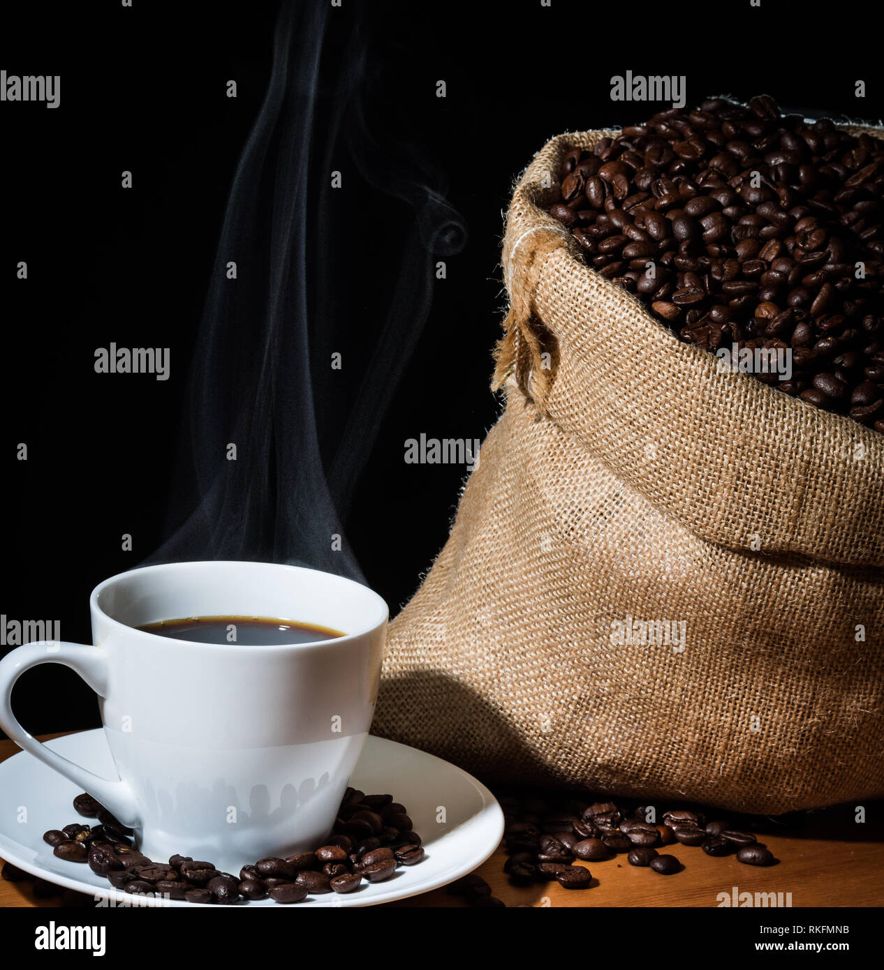 Gebrühten Kaffee in eine weiße Tasse mit Dampf oder Dunst und Kaffeebohnen in einem sackleinen hessische Sack und Bohnen auf einem hölzernen Fläche verstreut. Stockfoto