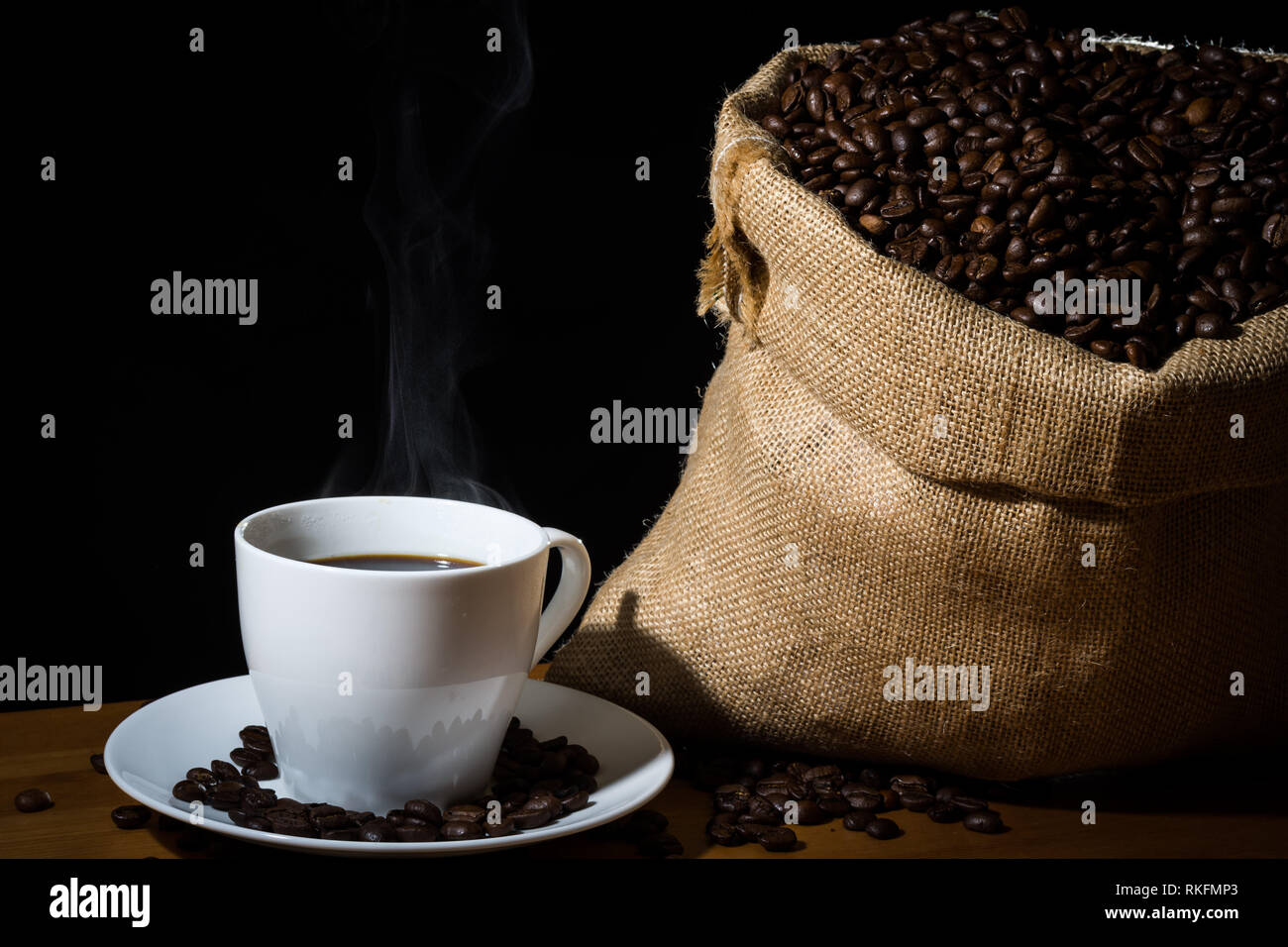 Gebrühten Kaffee in eine weiße Tasse mit Dampf oder Dunst und Kaffeebohnen in einem sackleinen hessische Sack und Bohnen auf einem hölzernen Fläche verstreut. Stockfoto