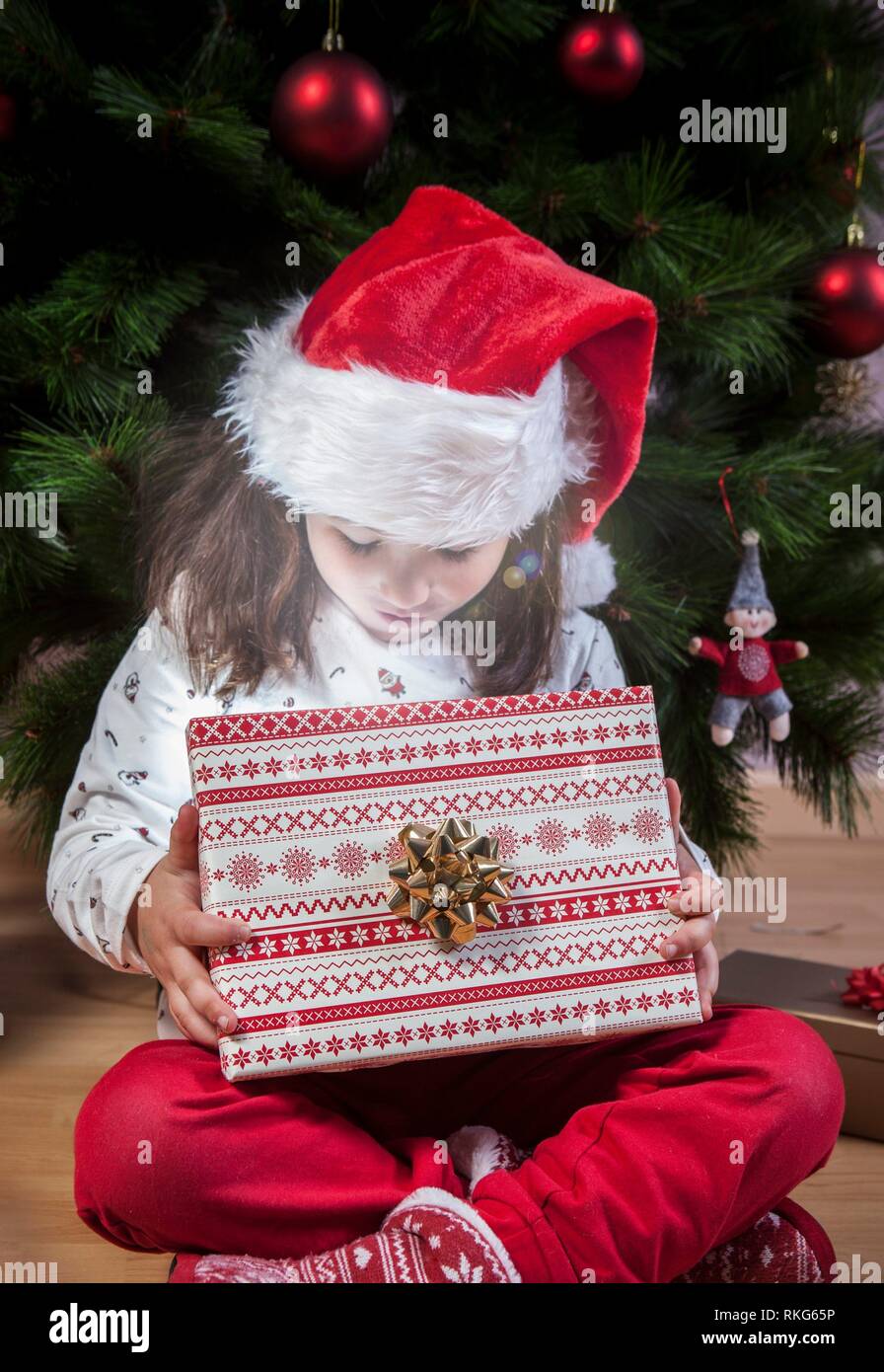 Kleines Mädchen öffnen ihre Gegenwart neben dem Weihnachtsbaum. Sie ist durch Glanz aus, beleuchtet. Stockfoto