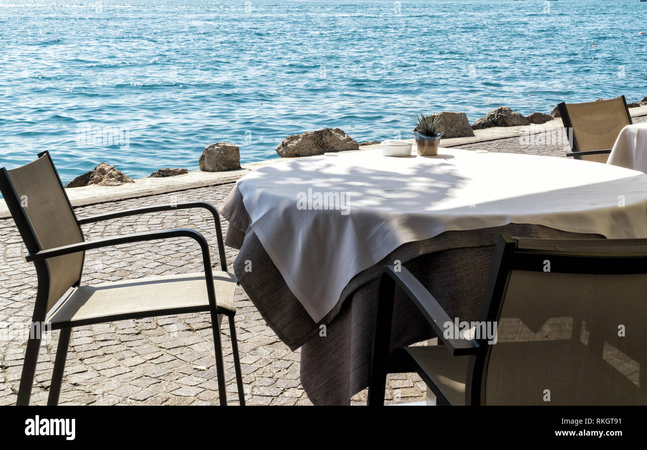 Leere Tabelle in einem Café am Ufer des Sees oder See, mit einer weißen Tischdecke mit Aschenbecher und einem dekorativen Blumentopf an einem sonnigen Sommertag abgedeckt. Stockfoto
