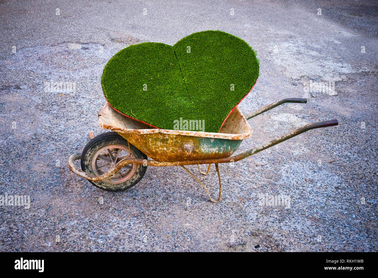 Ein Ausschnitt, der das Herz aus Kunststoff grün Gras an Bord, sitzt in einem alten benutzt Schubkarre auf ein uneinheitliches wenig Schotterstraße Stockfoto