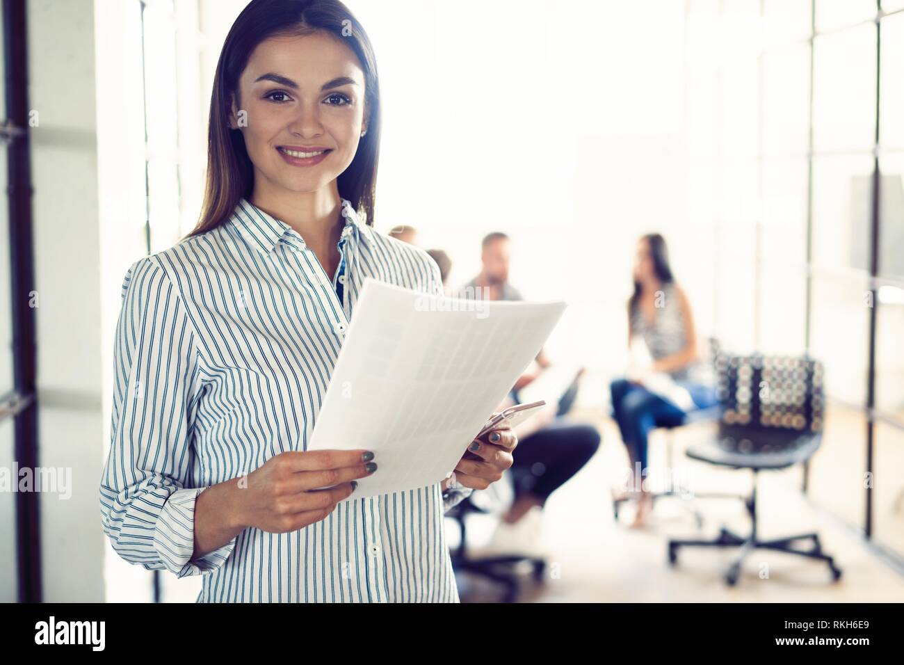 Selbstbewussten jungen Geschäftsfrau stehend mit Dokumenten, die in einem modernen Büro mit Kollegen im Hintergrund. Stockfoto