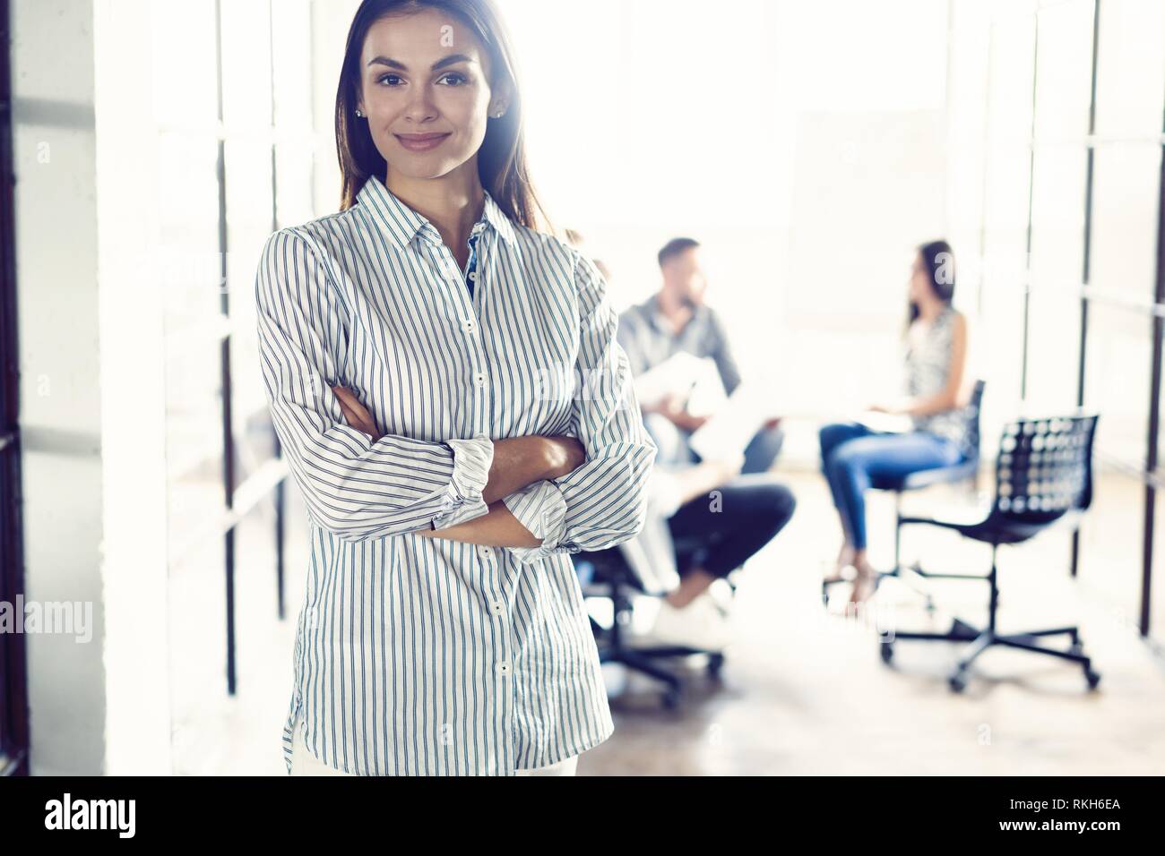 Selbstbewussten jungen Geschäftsfrau stehend, die Arme in einem modernen Büro mit den Kollegen im Hintergrund arbeiten gekreuzt. Stockfoto