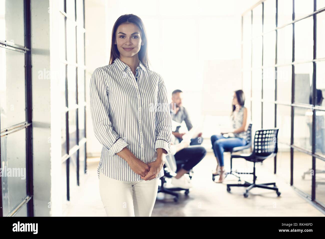 Selbstbewussten jungen Geschäftsfrau stehend, die Arme in einem modernen Büro mit den Kollegen im Hintergrund arbeiten gekreuzt. Stockfoto