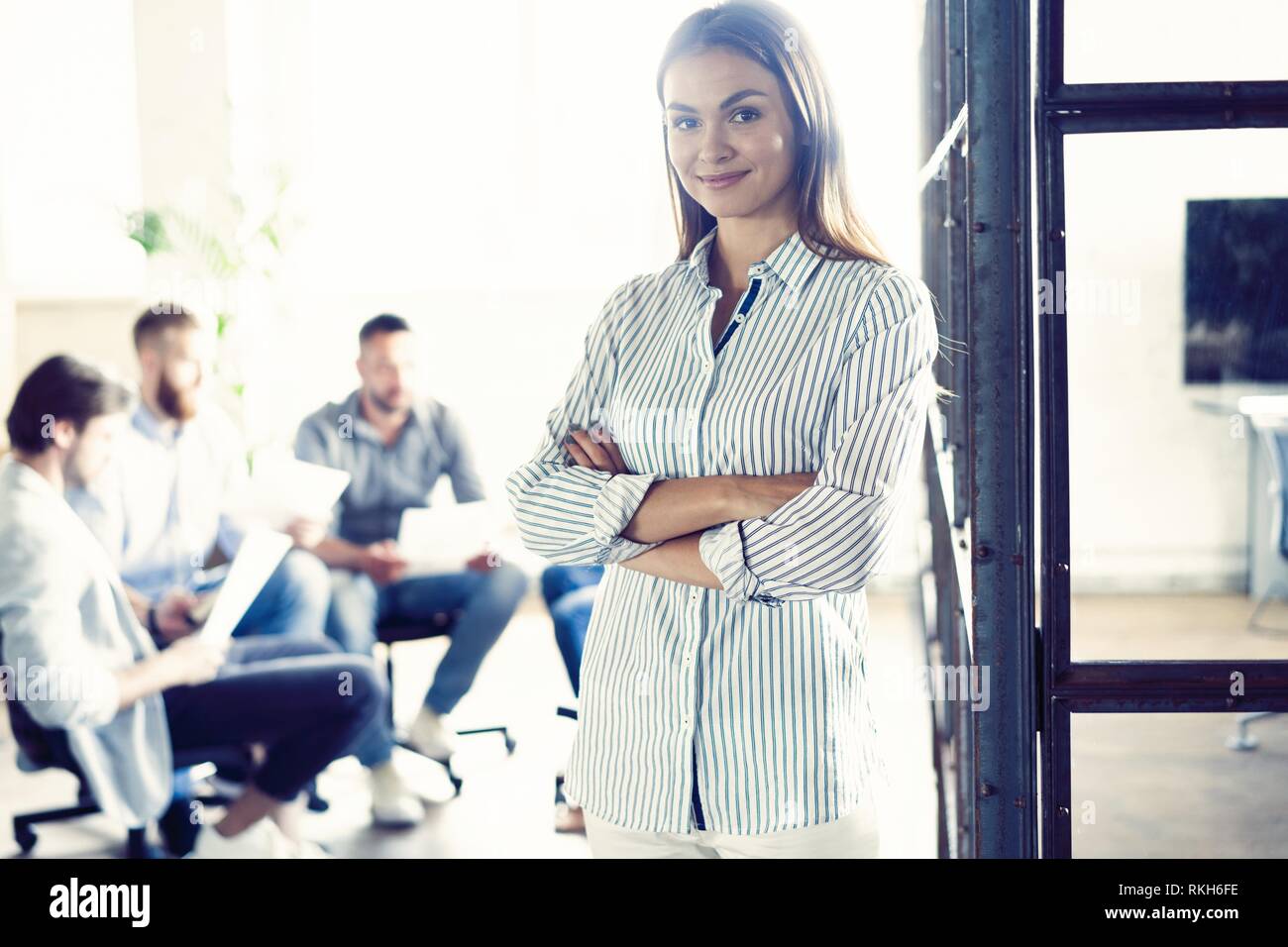 Selbstbewussten jungen Geschäftsfrau stehend, die Arme in einem modernen Büro mit den Kollegen im Hintergrund arbeiten gekreuzt. Stockfoto