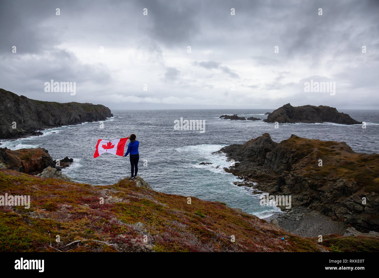 Abenteuerliche Frau mit einem kanadischen Flagge auf einem felsigen Atlantik Küste während eines bewölkten Tag. In sleepy Cove, Krähe Kopf, Twillingate, Newfoundl genommen Stockfoto