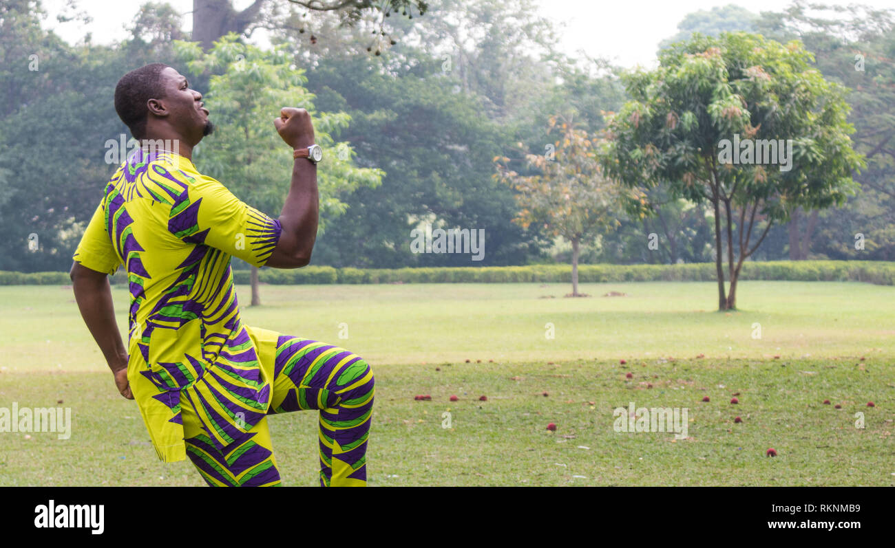 Ein afrikanischer Mann in ein offenes Feld des grünen Grases und klare Himmel punch die Luft und ein Bein in der Feier von einem Gewinn erhöhen. Stockfoto