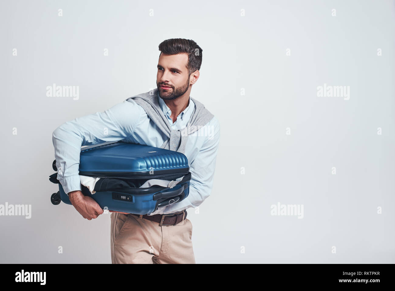 Bereit zu Reisen. Lächelnd schöner Mann hält Koffer voller Kleider beim Stehen auf einem grauen Hintergrund. Travel Concept. Close-up Portrait Stockfoto