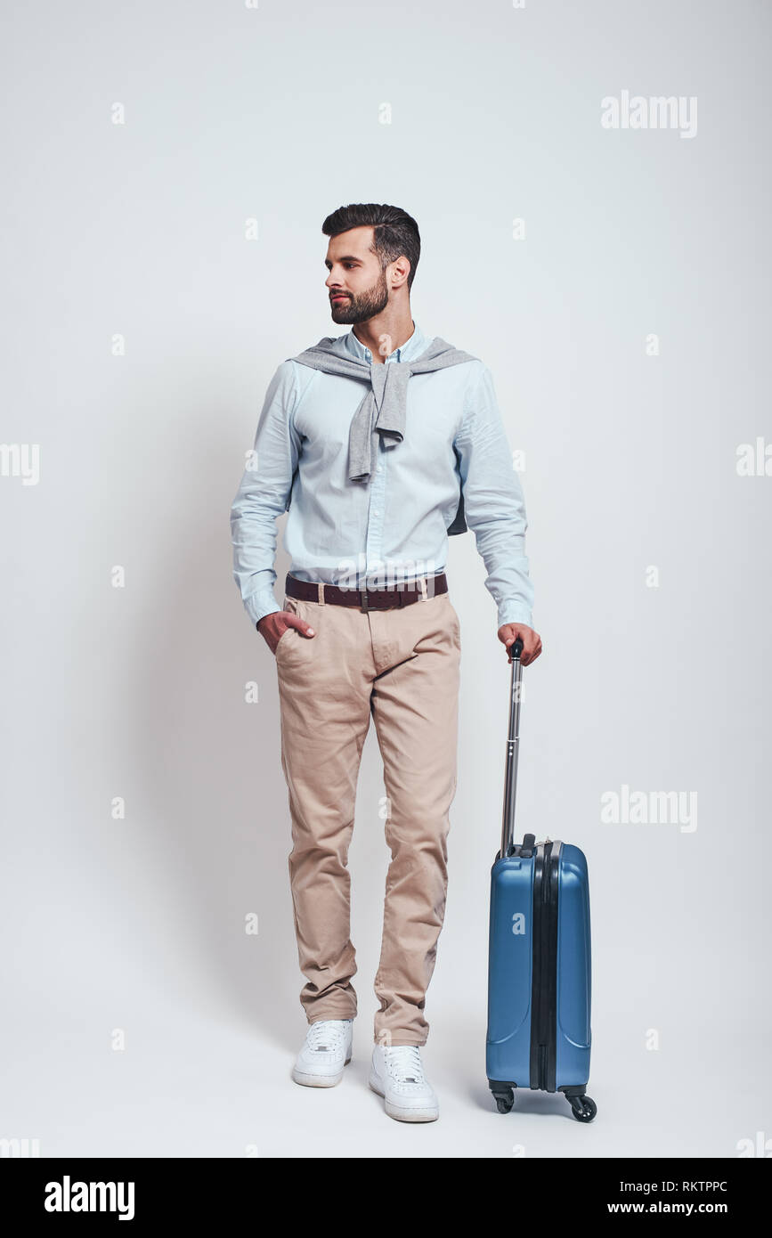 Bereit zu Reisen. Volle Länge von Stattlichen bärtiger junger Mann mit einem Koffer halten eine Hand in der Tasche weg schauen. Grauer Hintergrund. Travel Concept. Stockfoto