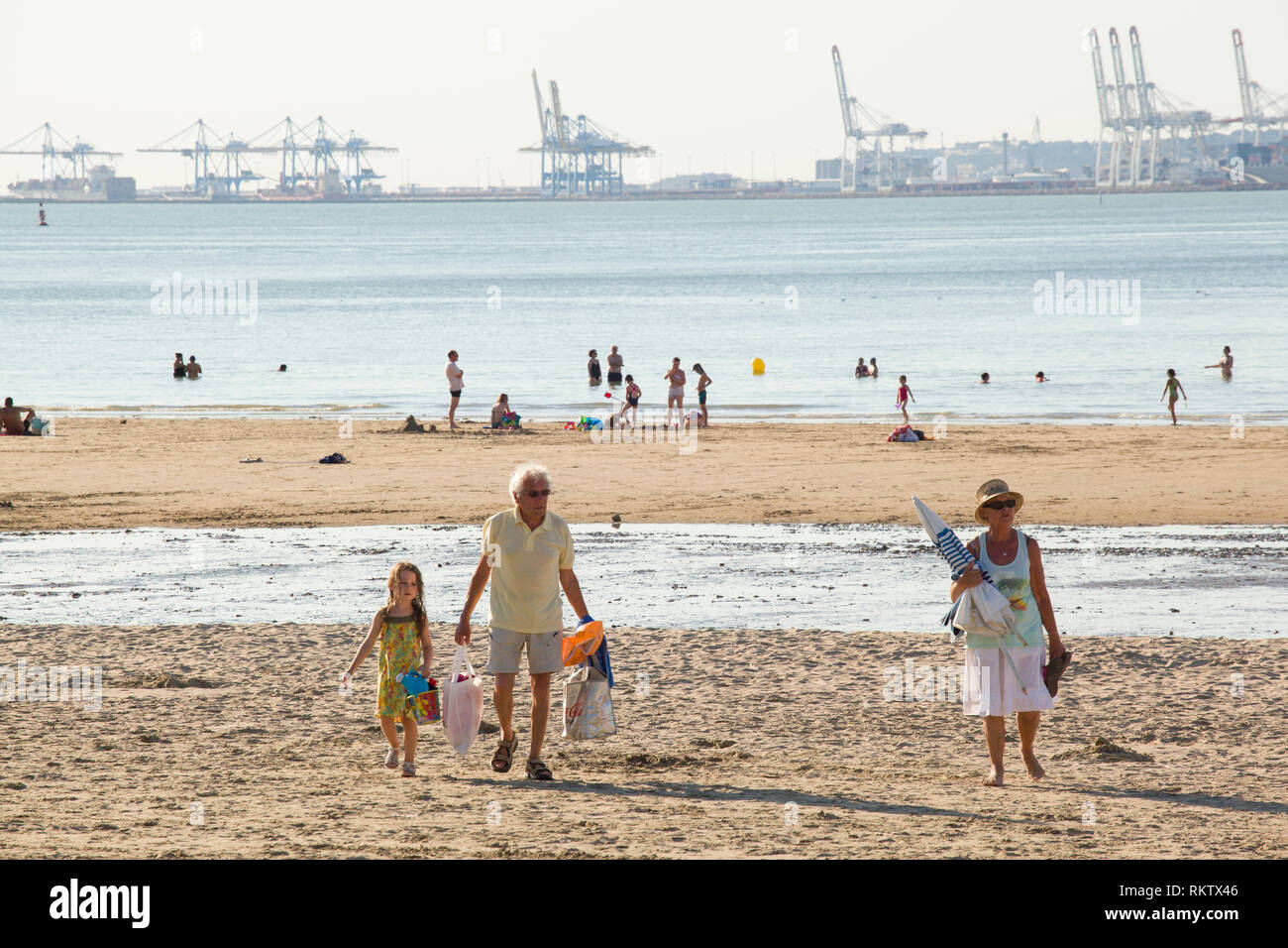 Urlauber genießen die Sonne Sommer am Strand Plage de Butin, Honfleur, Frankreich mit der Gantry Cranes Container Hafen von Le Havre über die Se Stockfoto