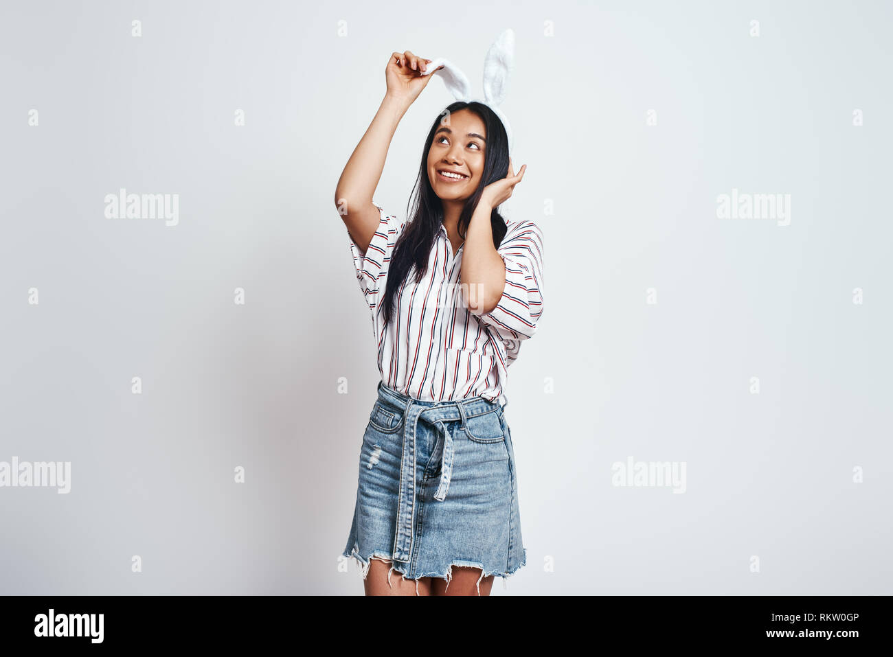 Tut, was sie will. Close up Portrait von jungen asiatischen Frau in legere Kleidung ihr bunny Ohren berühren und lächelnd. Grauer Hintergrund. Studio Portrait Stockfoto