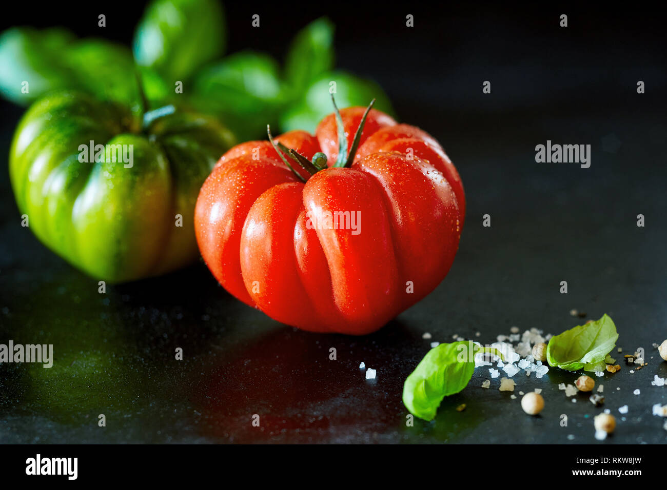 Frische ganze reife rote Tomaten mit Wassertropfen auf eine schwarze Fläche mit Kopie Raum und frischem Basilikum, grobes Salz und Pfeffer Stockfoto