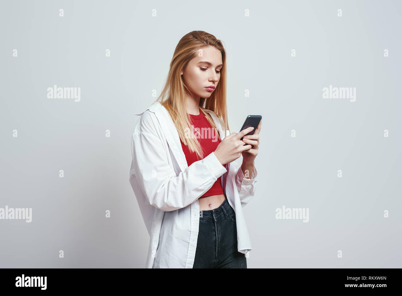 Immer verfügbar. Junge blonde Frau in einem Casual Wear mit Ihrem Smart Phone. Grauer Hintergrund. Digitale Konzept Stockfoto
