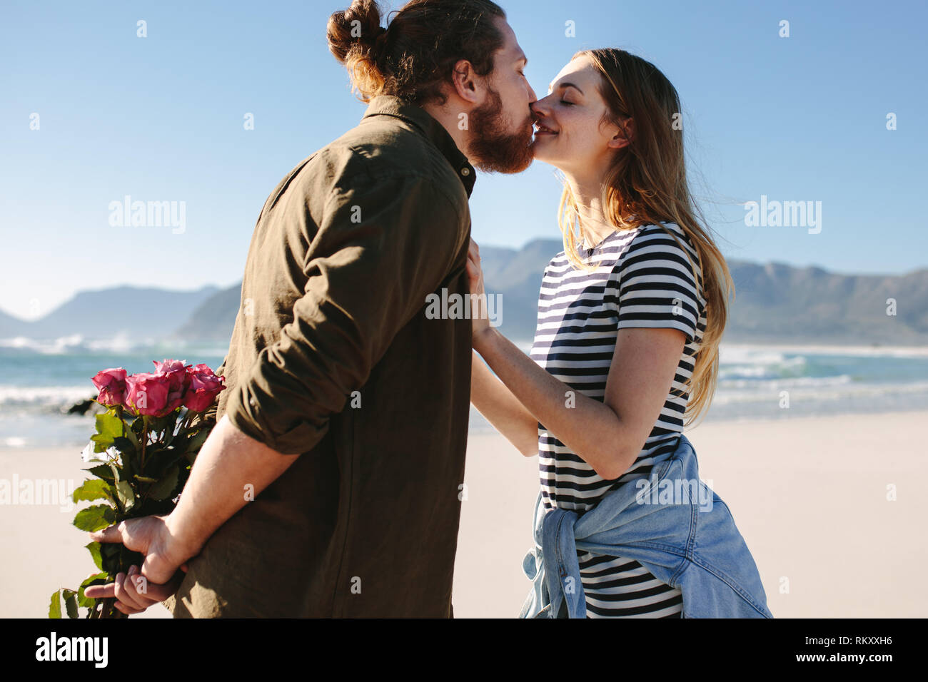 Mann mit Blumen hinter sich und küsste seine Freundin am Strand. Verliebtes paar Küssen auf den Strand. Stockfoto