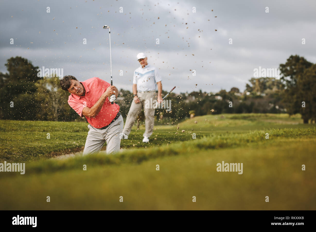 Ältere männliche Golfspieler Golf spielen auf dem Golfplatz. Der Mensch, der die Kugel aus einem Sandbunker mit seinem Gegner stehend an der Rückseite. Stockfoto
