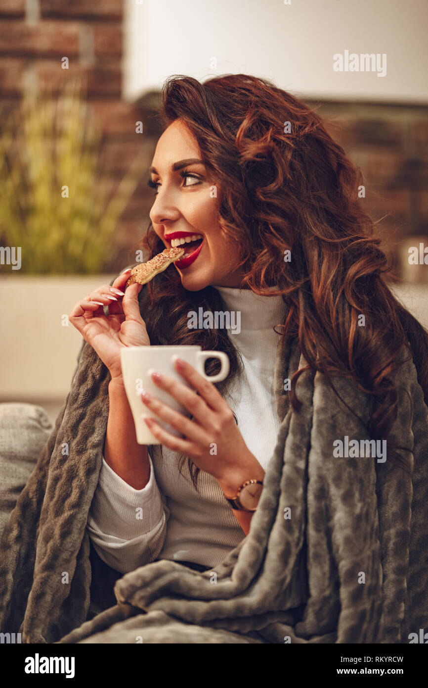 Eine schöne junge Frau sitzt auf dem Sofa in eine weiche Decke trinken Kaffee am Morgen verstaut und beißt Cookie. Stockfoto
