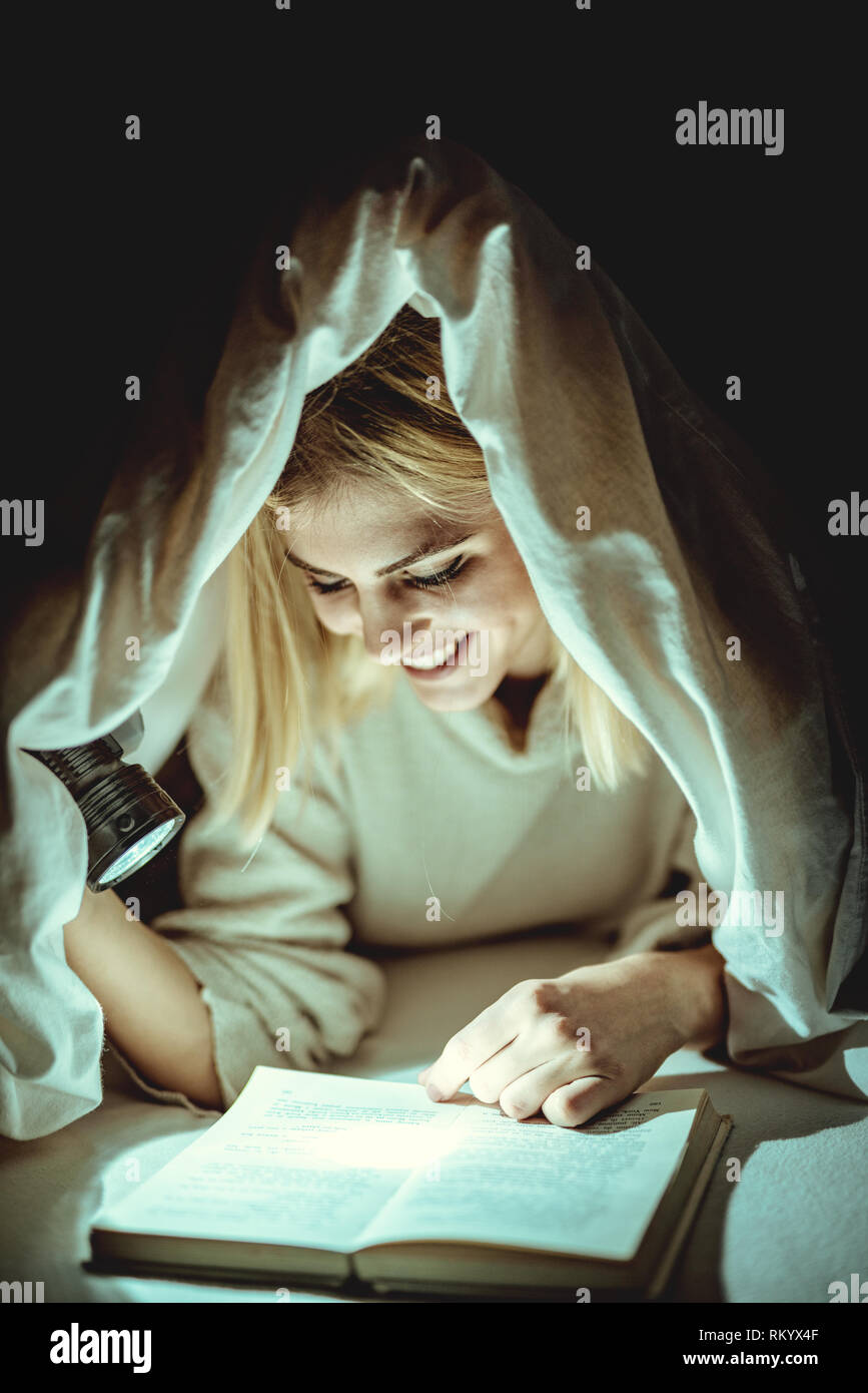 Schöne junge Frau liest Buch unter dem Bett auf und hält eine Taschenlampe. Stockfoto