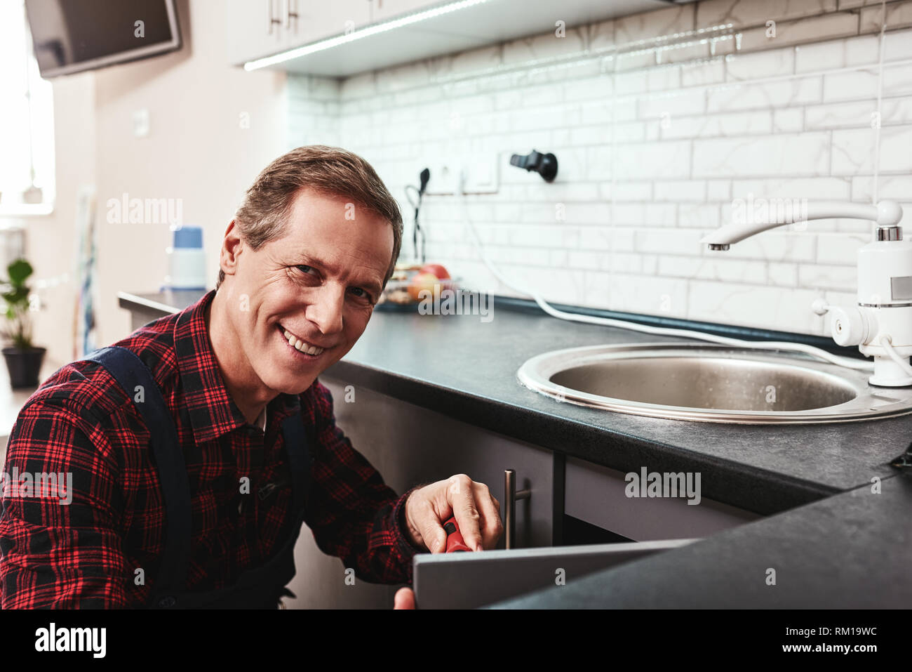 Lächelnd Master. Seniour Heimwerker Instandsetzung Waschbecken in der Küche mit einem Schraubendreher Stockfoto